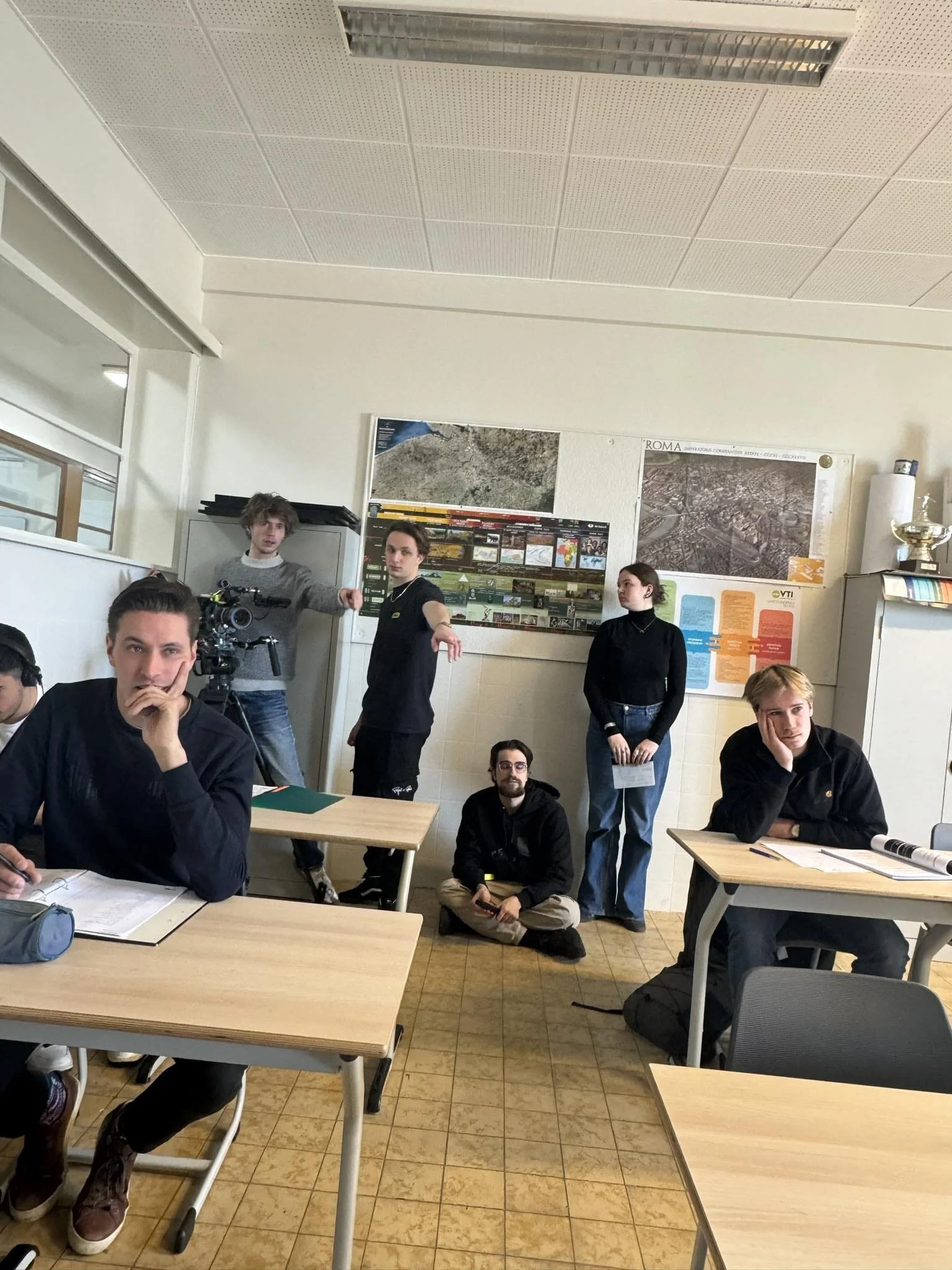 Group of students in a classroom, some sitting at desks and others standing, with a camera on a tripod in the background, and posters on the wall.