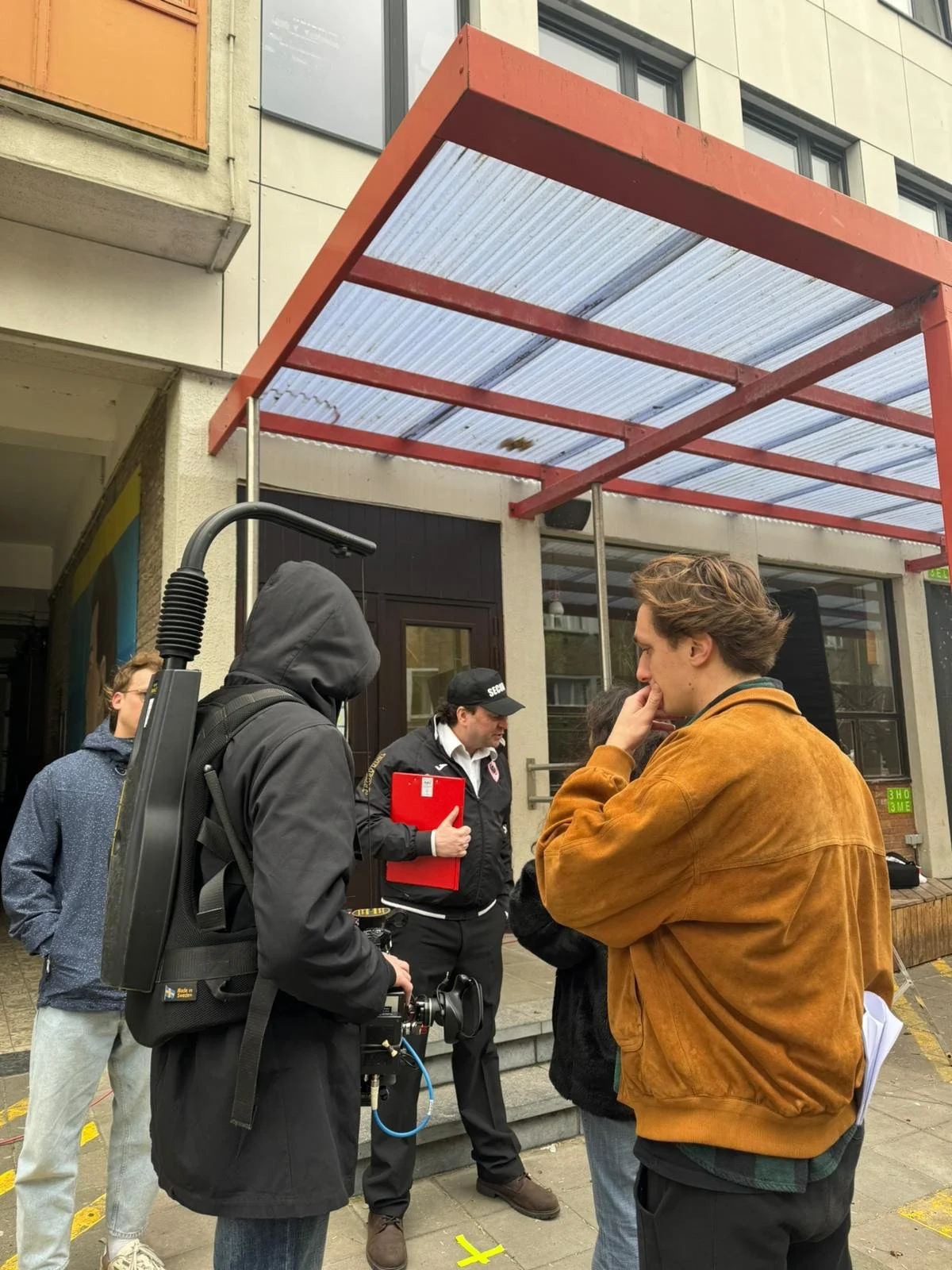 A group of young people and a man with a red folder standing outside a building, engaged in a discussion, with some equipment and a camera visible.