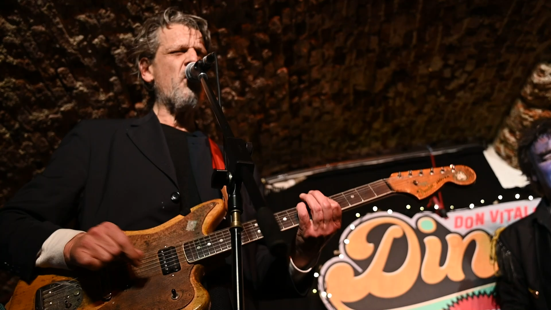 A bearded man with gray hair, wearing a black blazer and dark shirt, is playing an electric guitar and singing into a microphone. There is a colorful sign with the word "Dun" and a red and white sign that reads "Don Vital" in the background, with a stone ceiling above.