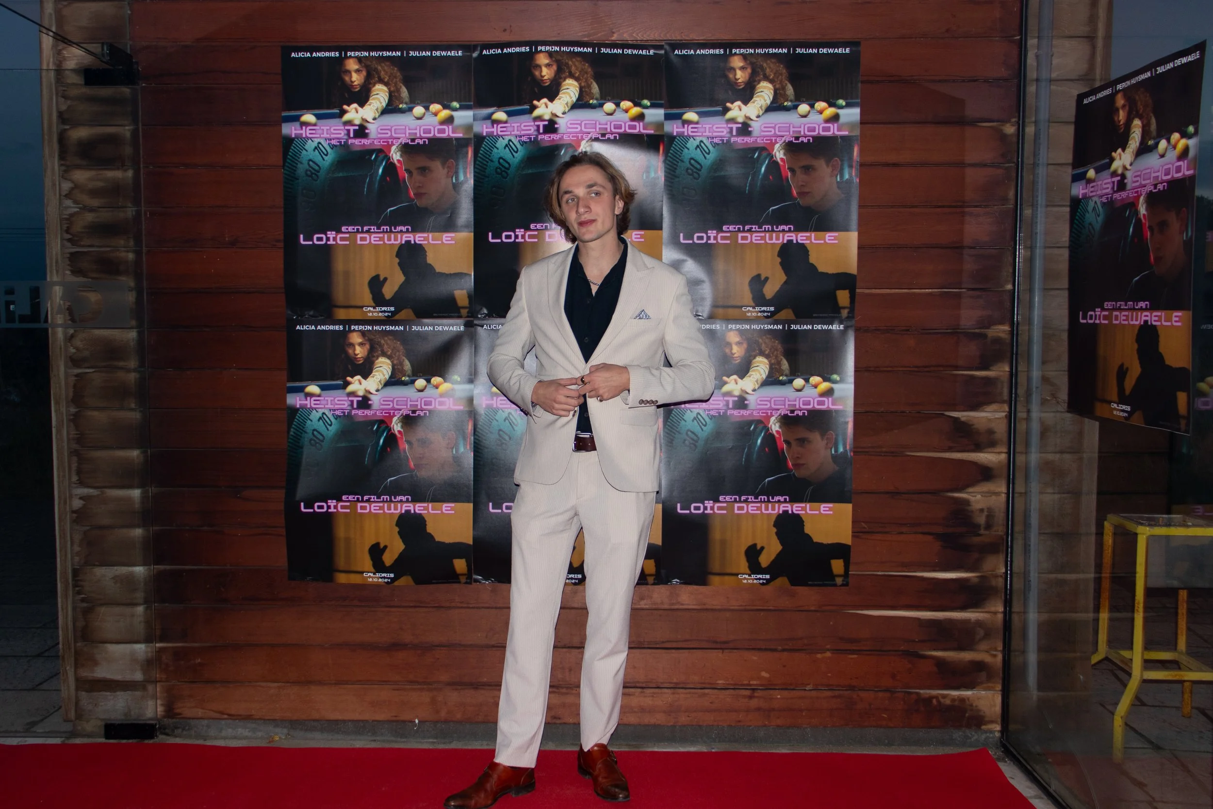 A young man in a white suit standing on a red carpet in front of a wooden wall with movie posters for 'Heist School' featuring images of a woman playing pool and a man with a serious expression.