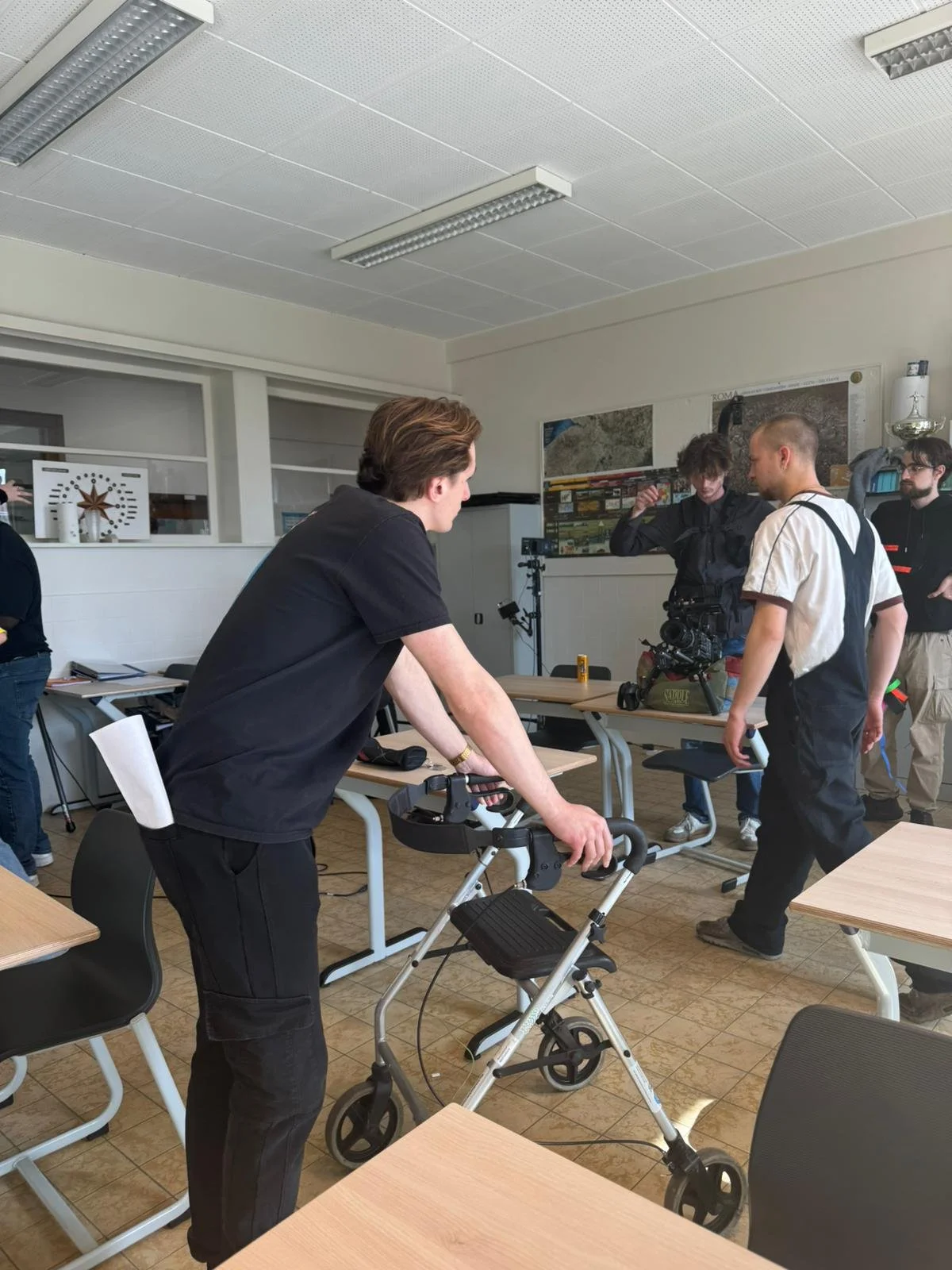 A group of people in a classroom setting, with one person using a walker and others engaging with a camera. The room has a white ceiling with fluorescent lights, desks, and chairs, and posters on the wall.