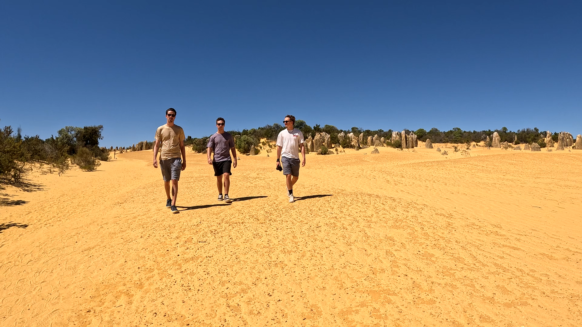 Three young men walking in a sandy desert with sparse vegetation and rock formations under a clear blue sky.