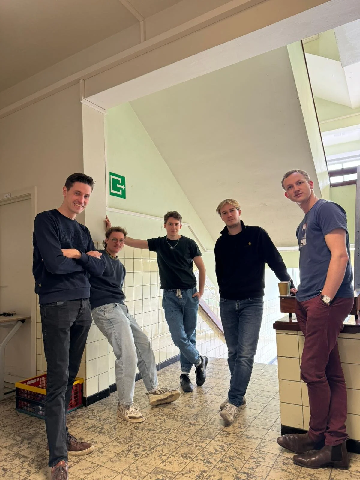 Five young men standing in a corner of a tiled hallway with a staircase behind them. They are casually dressed and posing for the photo.