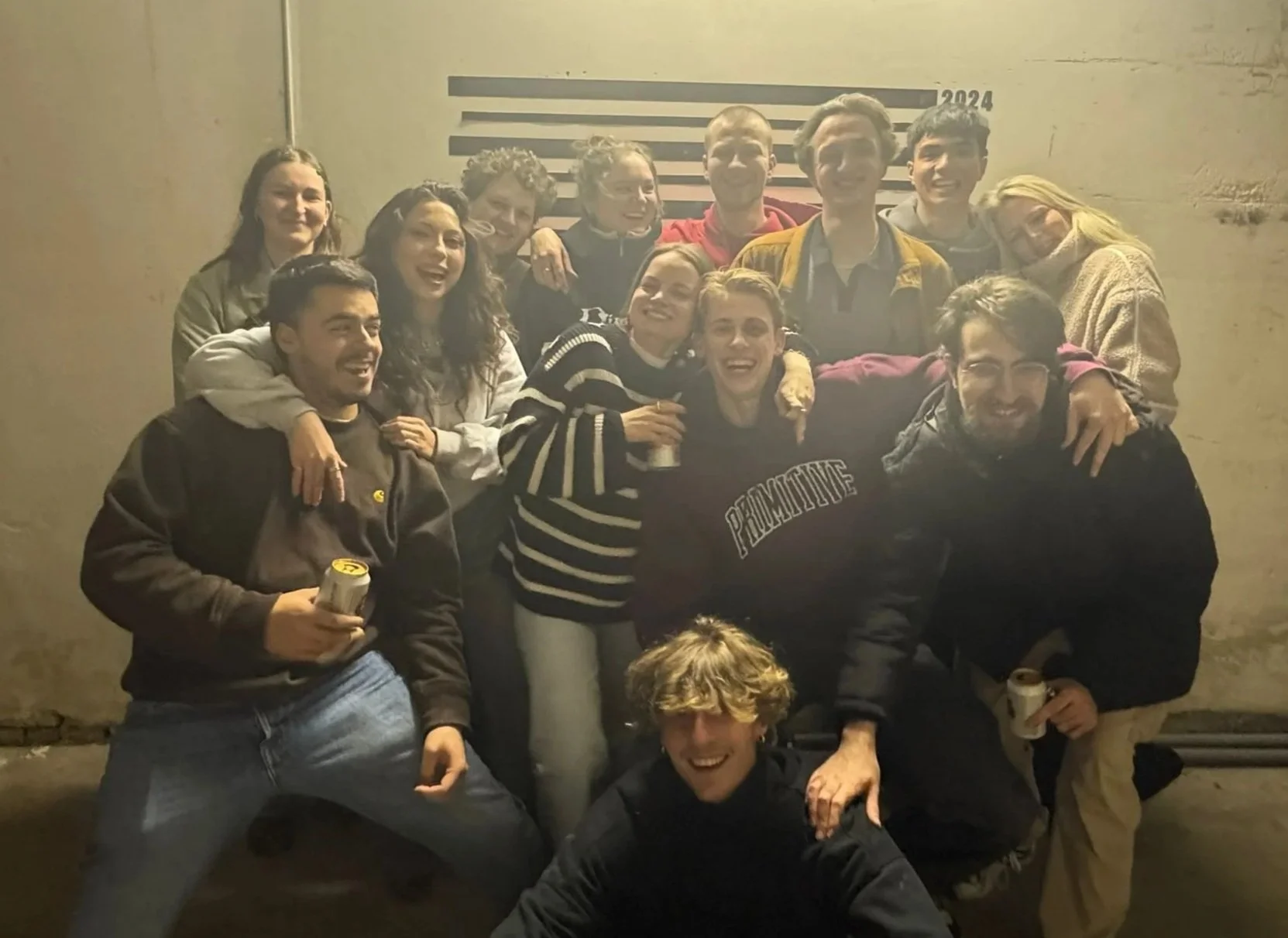 Group of young adults smiling and celebrating together, some holding drinks, in an industrial setting with a height measurement chart in the background.