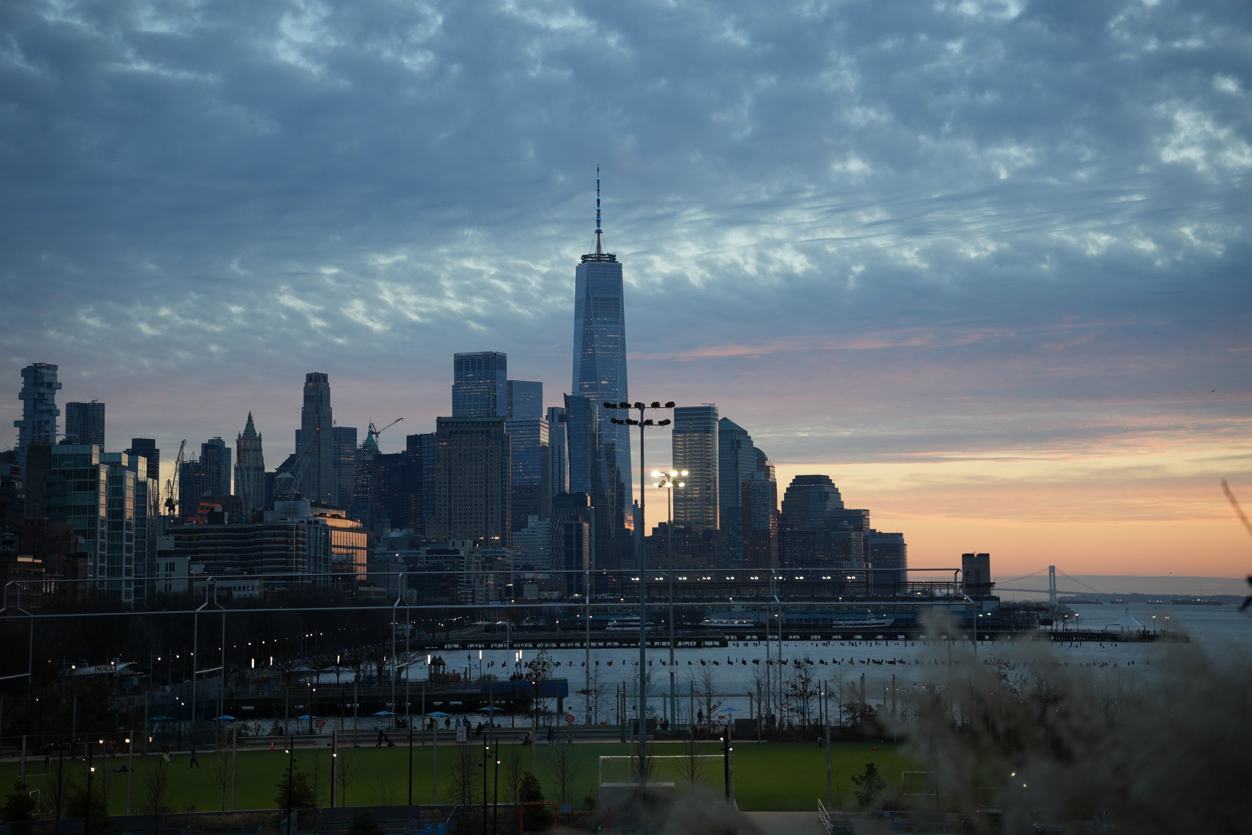 City skyline of New York City at dusk with skyscrapers, including One World Trade Center, and a river in the foreground.