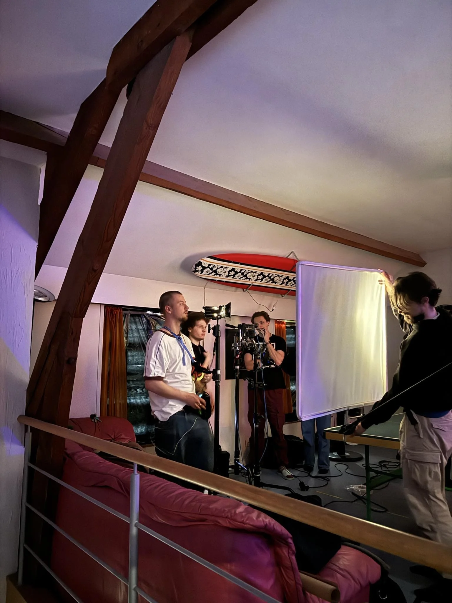 A behind-the-scenes shot of a film or photo shoot setup in a cozy room with wood beams. Several crew members are gathered around a camera, with one adjusting a large reflector or diffuser, and others monitoring the scene. A skateboard with a floral d