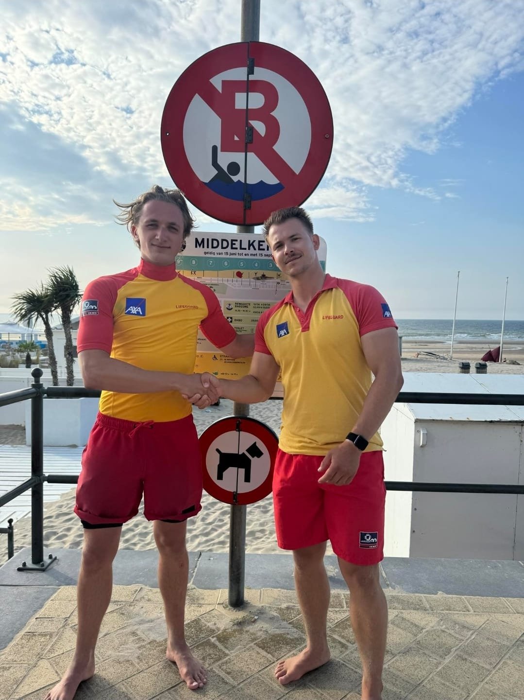 Two lifeguards in yellow and red uniforms shaking hands at the beach, with signs indicating no swimming and no dogs, and the ocean in the background.