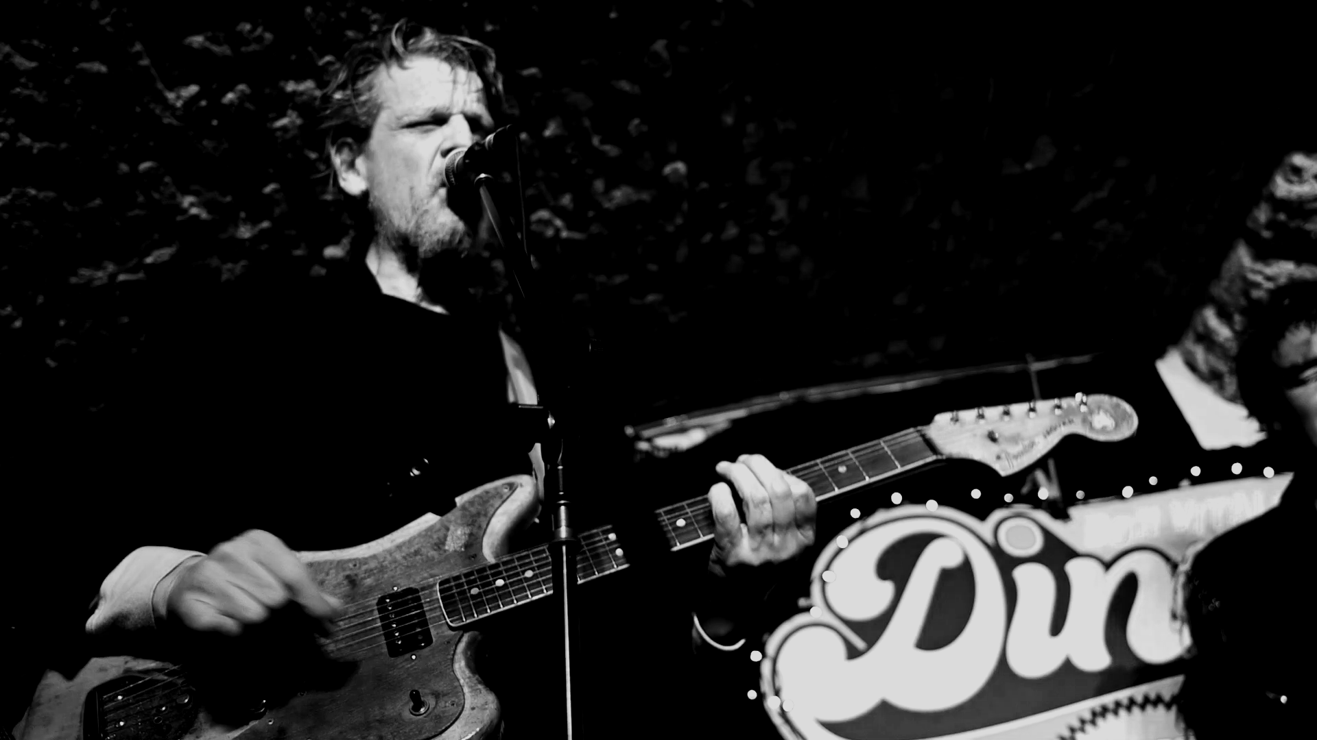 A man with long hair and a beard passionately singing into a microphone while playing an electric guitar during a performance, with a partially visible sign reading 'Din' in the background.