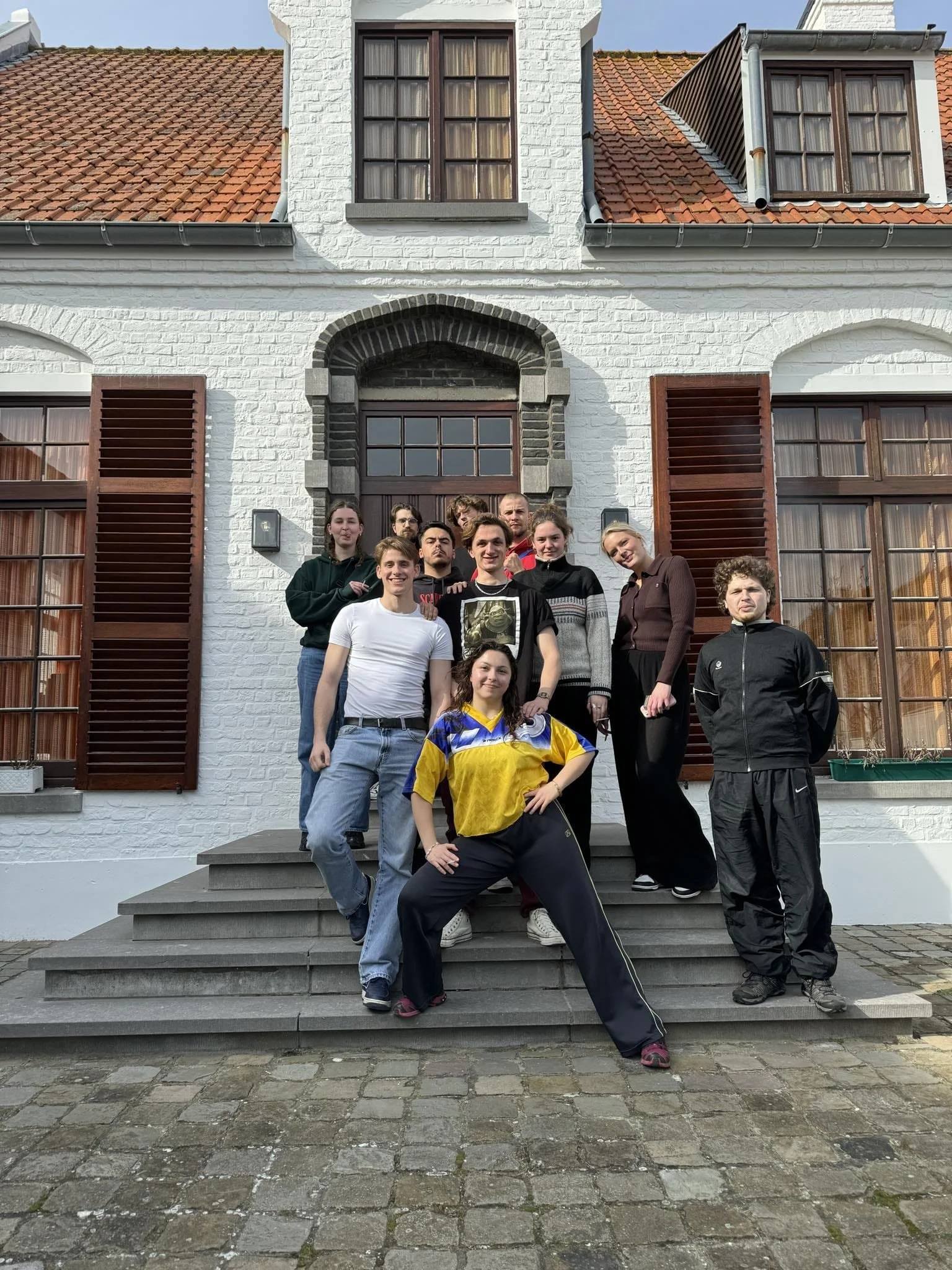 Group of ten young people standing on a set of outdoor stairs in front of a white brick house with brown shutters and a red-tiled roof.