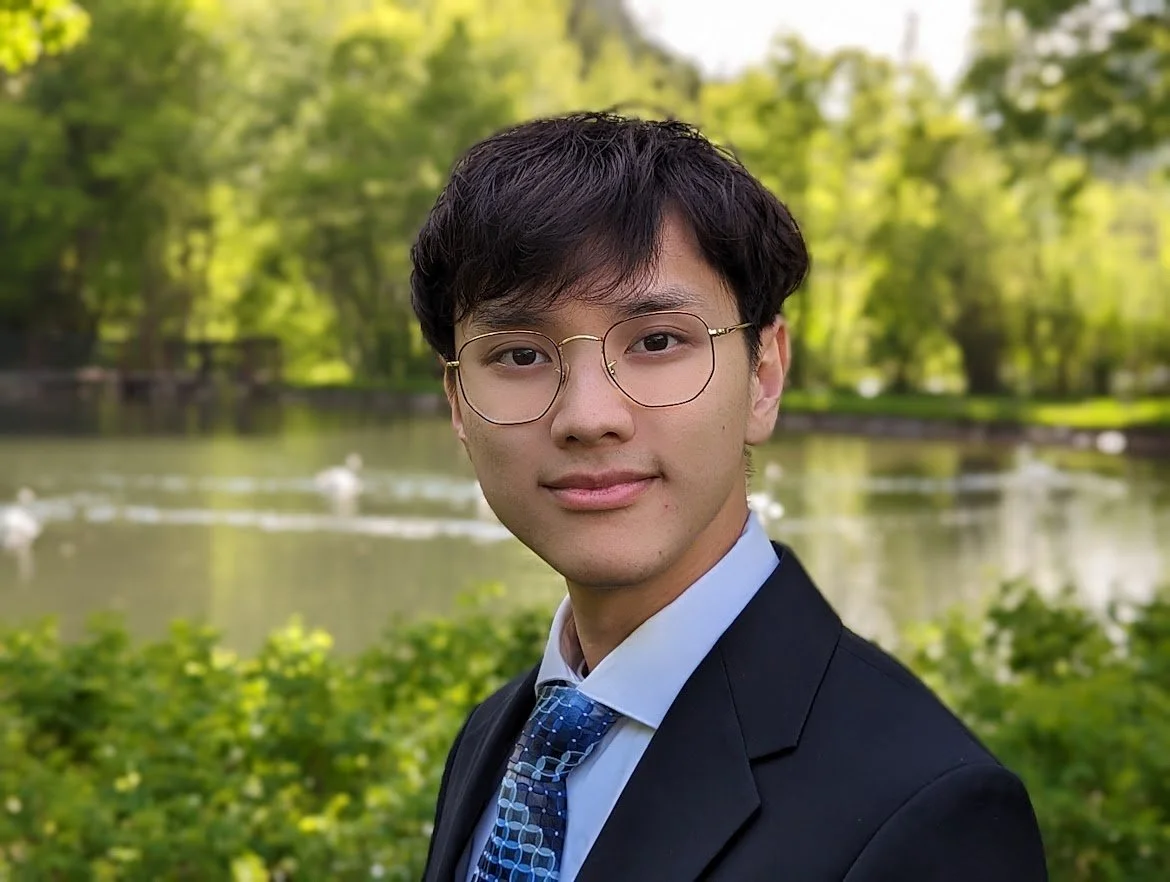 A young man in a suit with a blue tie and glasses, standing outdoors near a pond with ducks, surrounded by green trees.