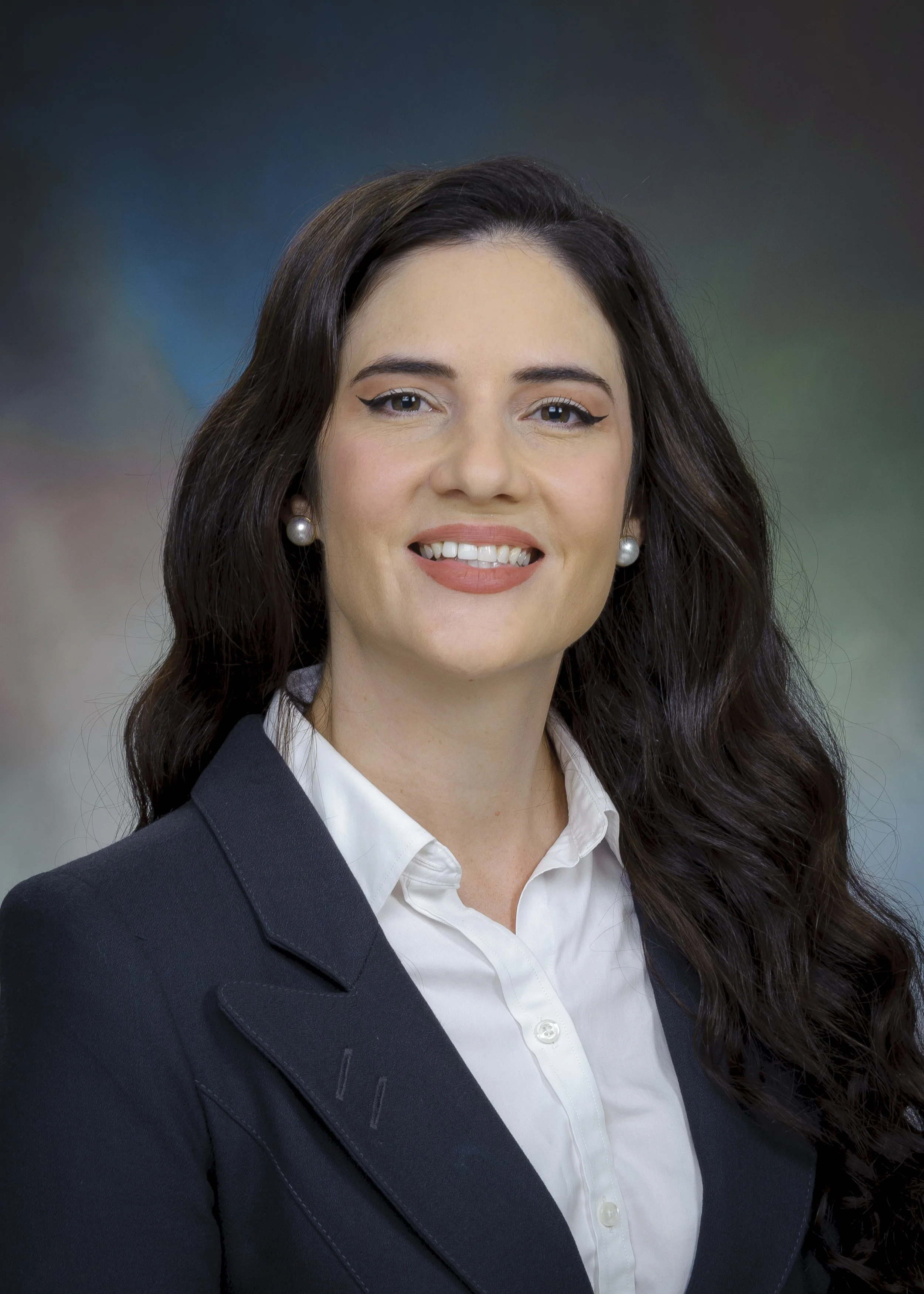 A professional woman with long dark hair, wearing a black blazer, white shirt, pearl earrings, and makeup, smiling at the camera with a colorful blurred background.