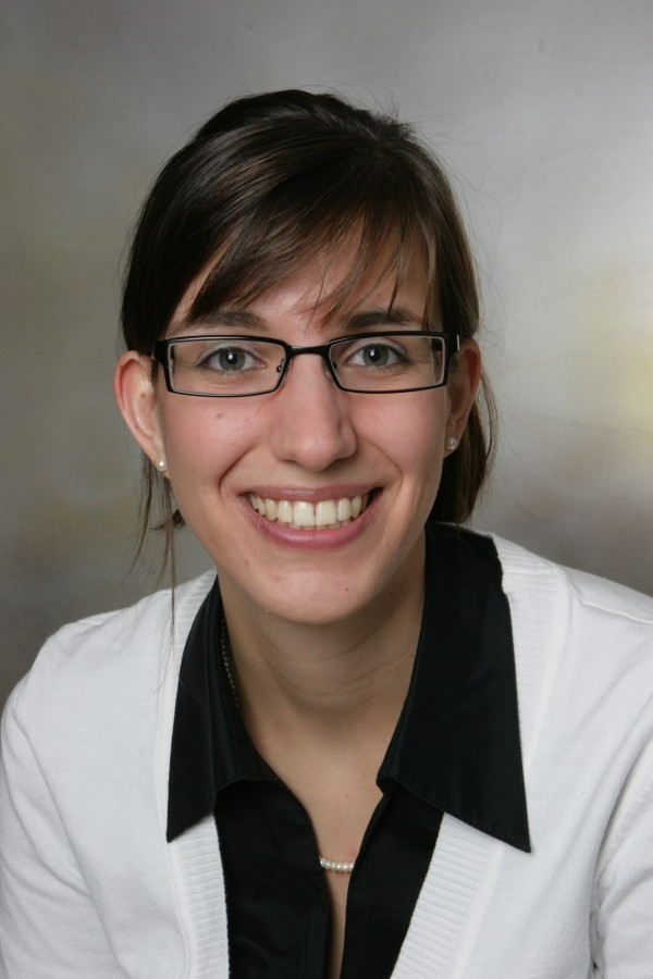 A woman with dark brown hair, glasses, and earrings smiling in a professional setting, wearing a black shirt and white cardigan.