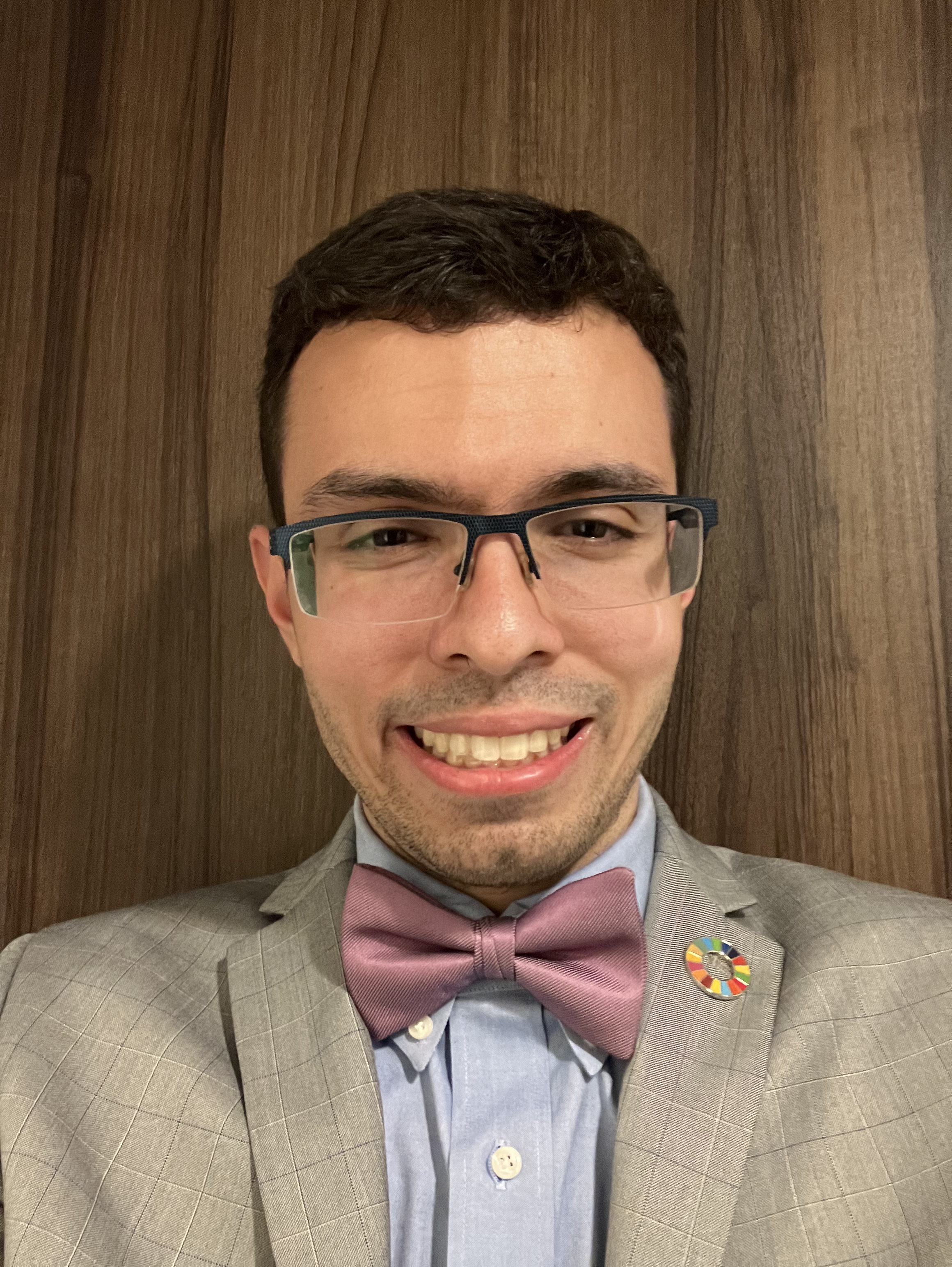 A young man in a gray suit, pink bow tie, and glasses taking a selfie in front of a wooden wall, wearing a circular pin with colorful segments.