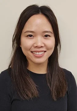 A woman with long dark hair smiling and wearing a black top against a light background.