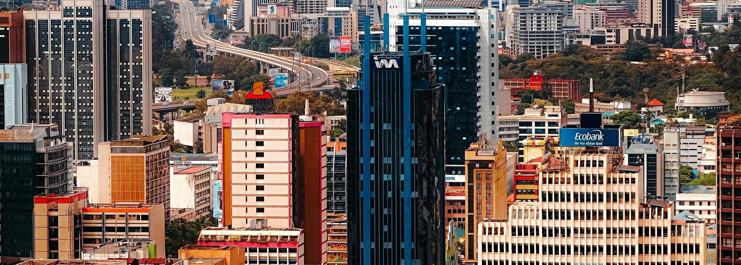 A city skyline with tall skyscrapers, some under construction, and a highway curving through the city. Various buildings have signs, including one for Ecobank and WM.
