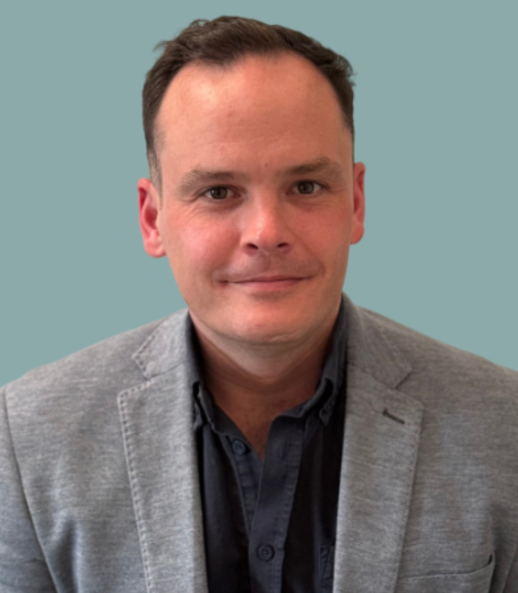 A photo of Kieran, gray blazer and a dark button-up shirt, against a plain teal background.