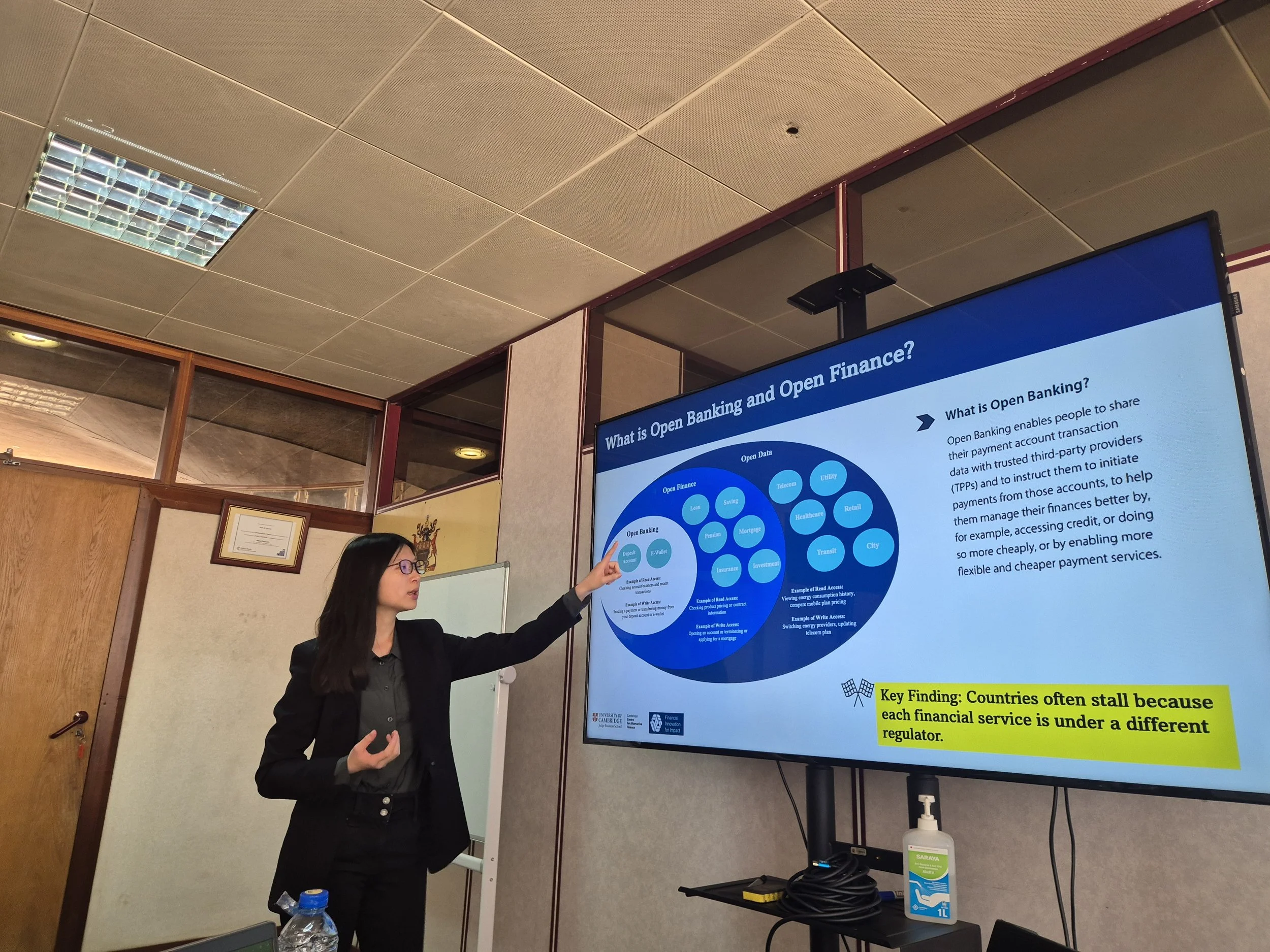 A woman in business attire and glasses is giving a presentation in front of a large screen. The screen displays a slide about open banking and open finance, with various diagrams and text.