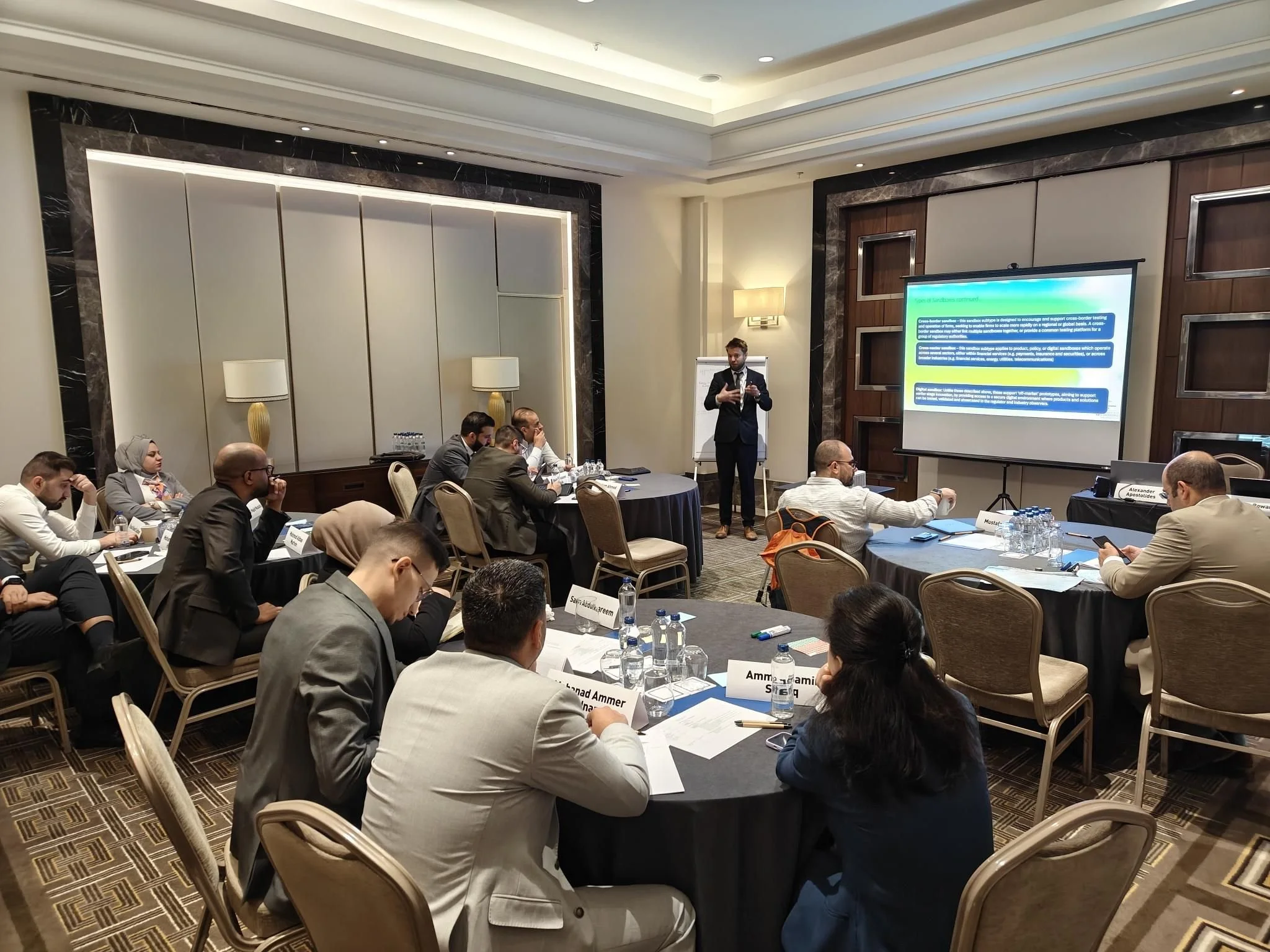 Business conference room with attendees seated around tables, a presenter in a suit standing at the front near a large screen displaying a PowerPoint presentation.