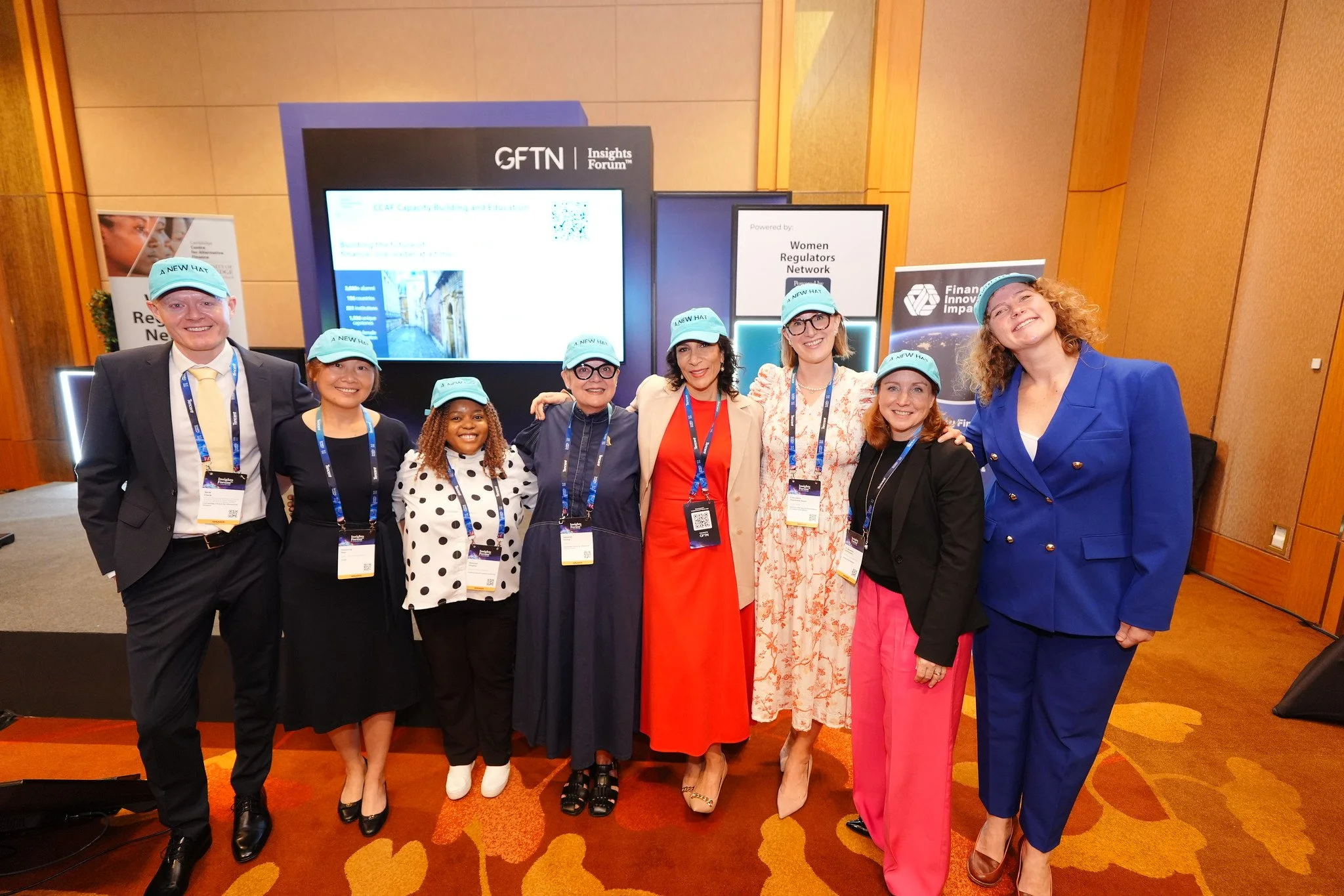 Group of eight diverse professionals standing together at a conference, all wearing matching light blue hats and smiling for the camera.
