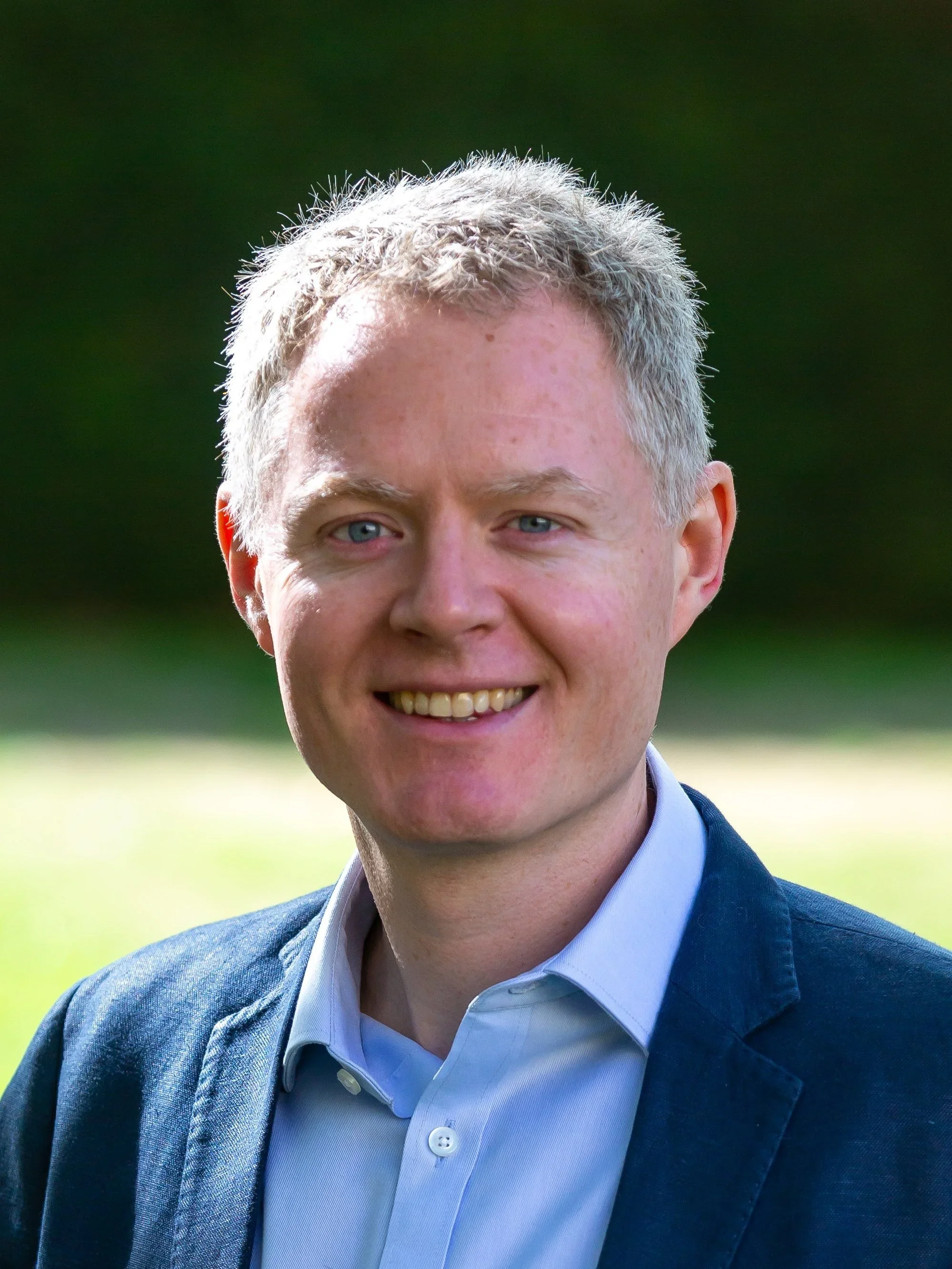 A photo of Nick, a man wearing a light blue collared shirt and a dark blue blazer, outdoors with a blurred green background.