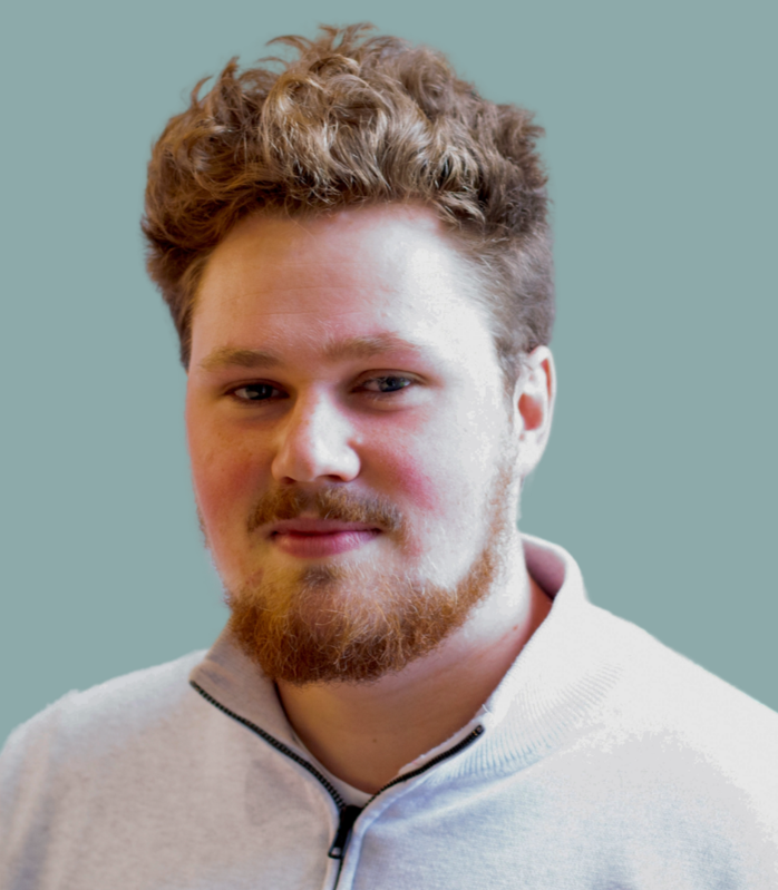 A young man with curly brown hair and a beard, wearing a light-colored zip-up sweater, looking at the camera against a plain background.
