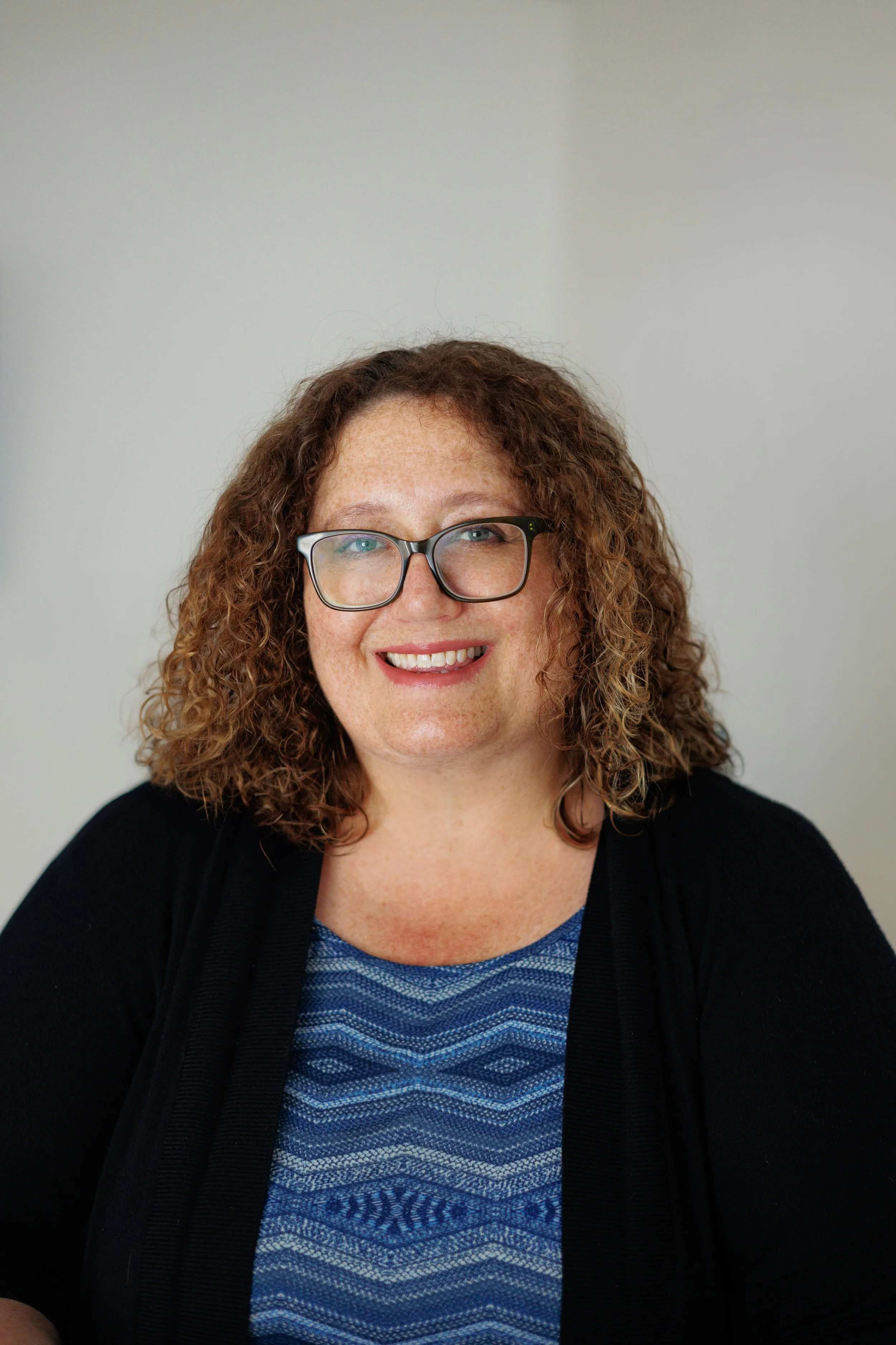 A woman with curly hair, glasses, and a blue patterned top, smiling at the camera against a plain background.