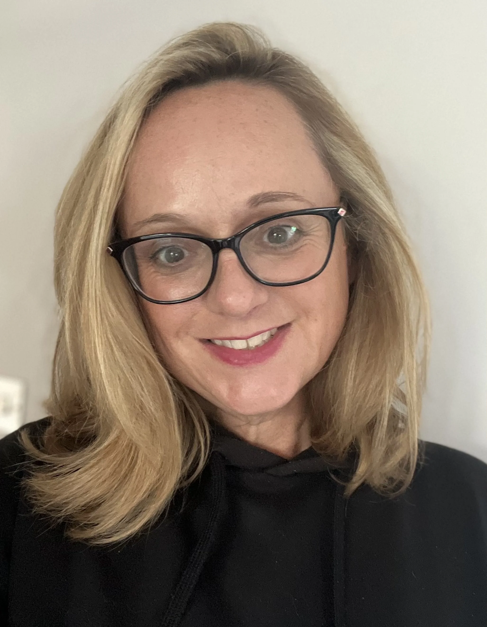 Close-up of a smiling woman with shoulder-length blonde hair, wearing black glasses and a black hoodie, standing against a light-colored wall.