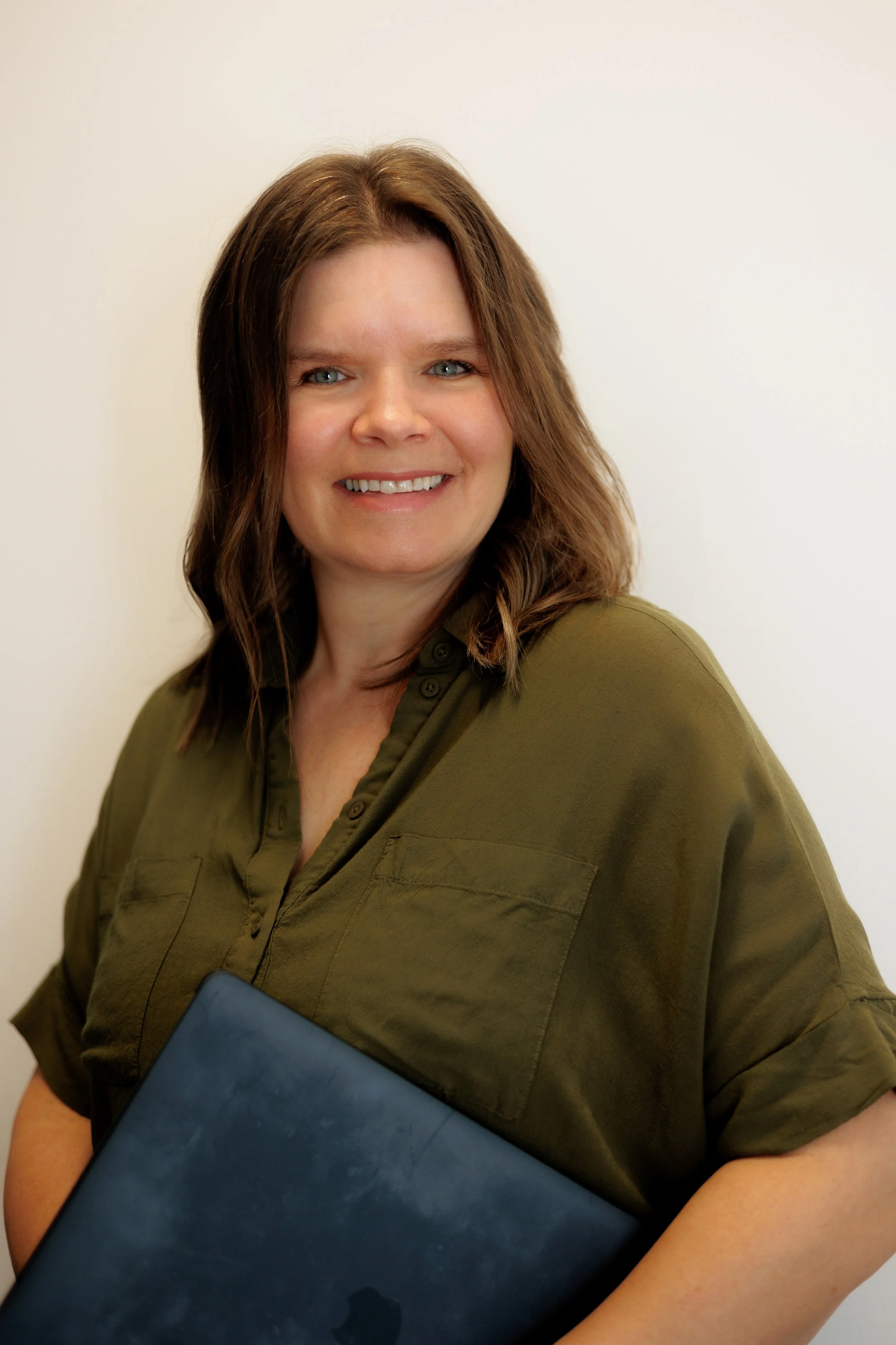 A woman with shoulder-length brown hair, wearing a green button-up shirt, smiling and holding a blue laptop.