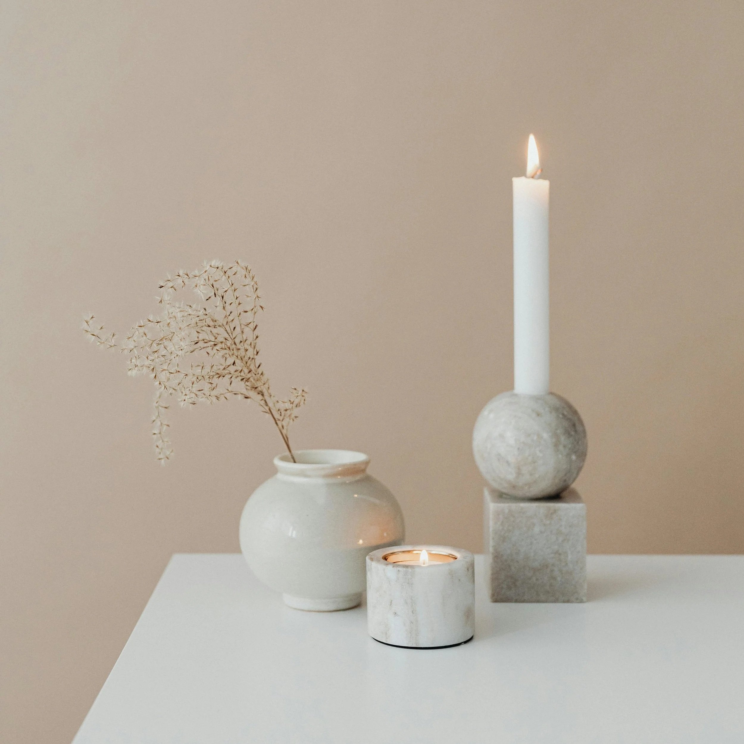 Minimalist white table with a ceramic vase holding dried flowers, a short candle in a marble holder, and two tall candles in cube and sphere holders against a neutral background.
