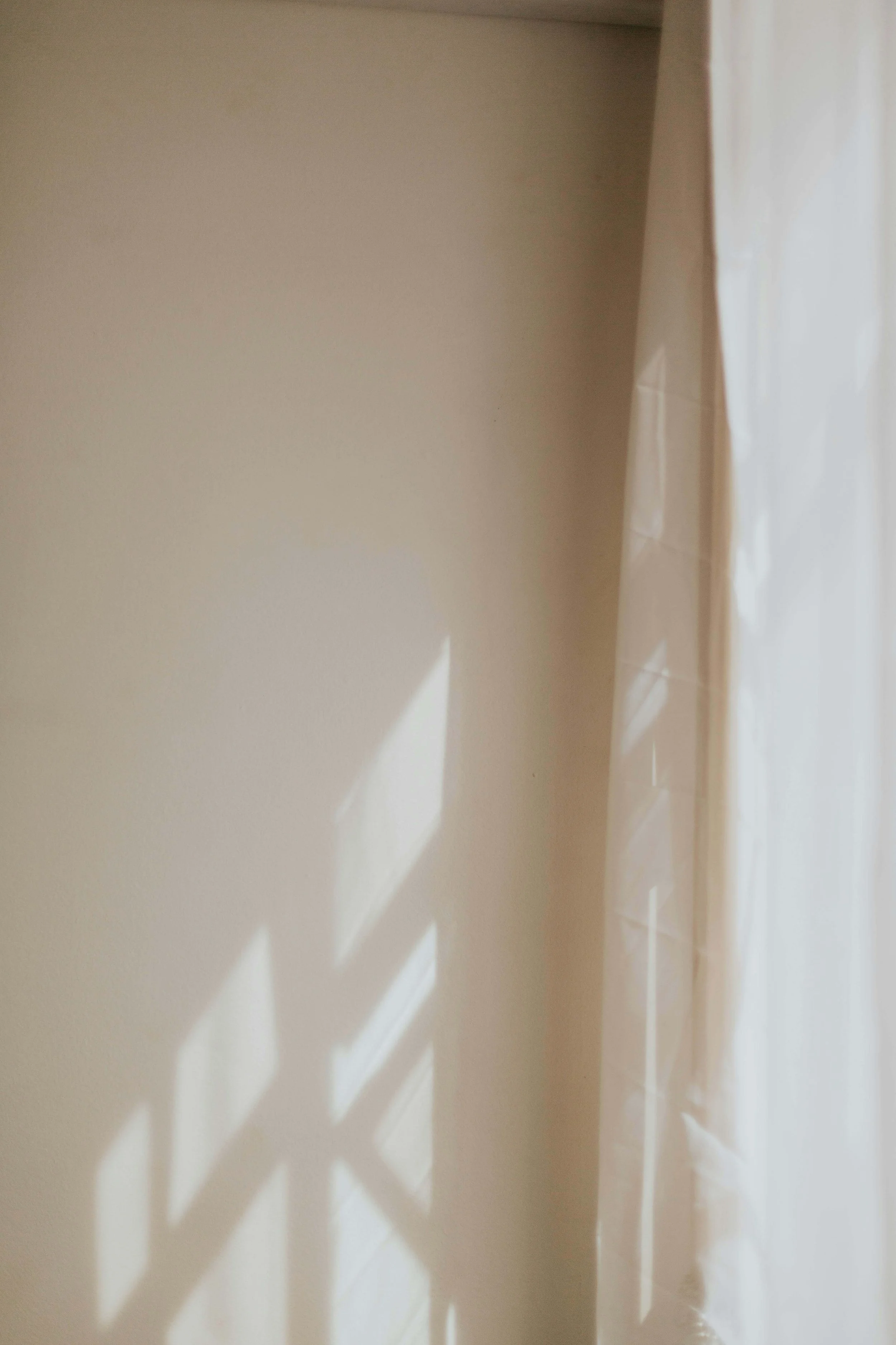 Sunlight streaming through a window creates geometric shadow patterns on a beige wall and white curtain.