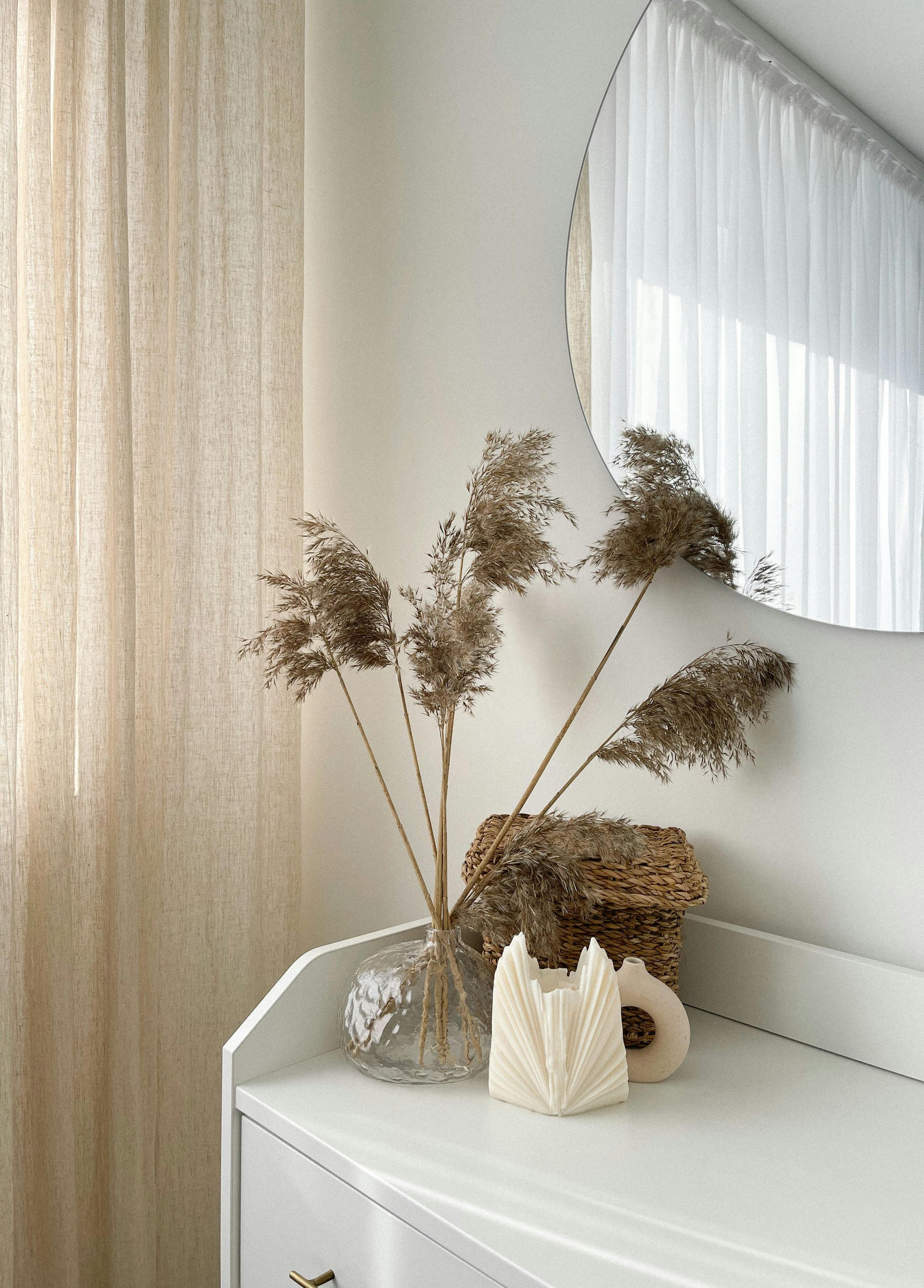 Decorative wooden dresser with a clear glass vase holding dried pampas grass, a woven basket, a white ceramic vase, and a white decorative sculpture, next to a large round mirror on a white wall.