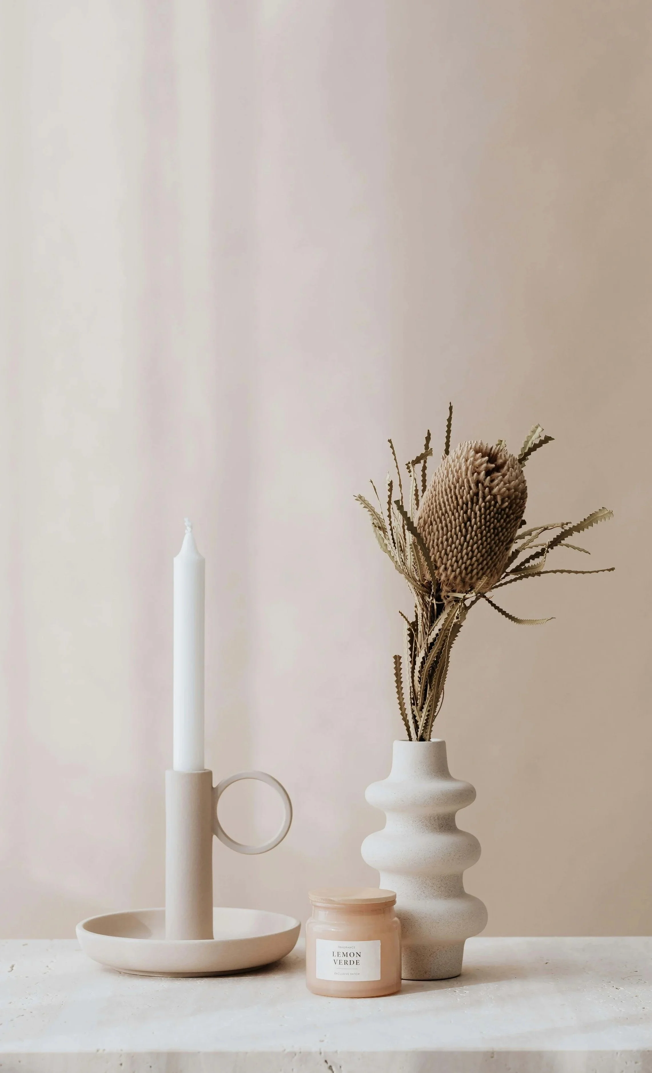 A beige candle in a holder, a white vase with dried flowers, and a pink candle with a label reading 'Lemon Verde' on a light-colored surface against a plain wall.