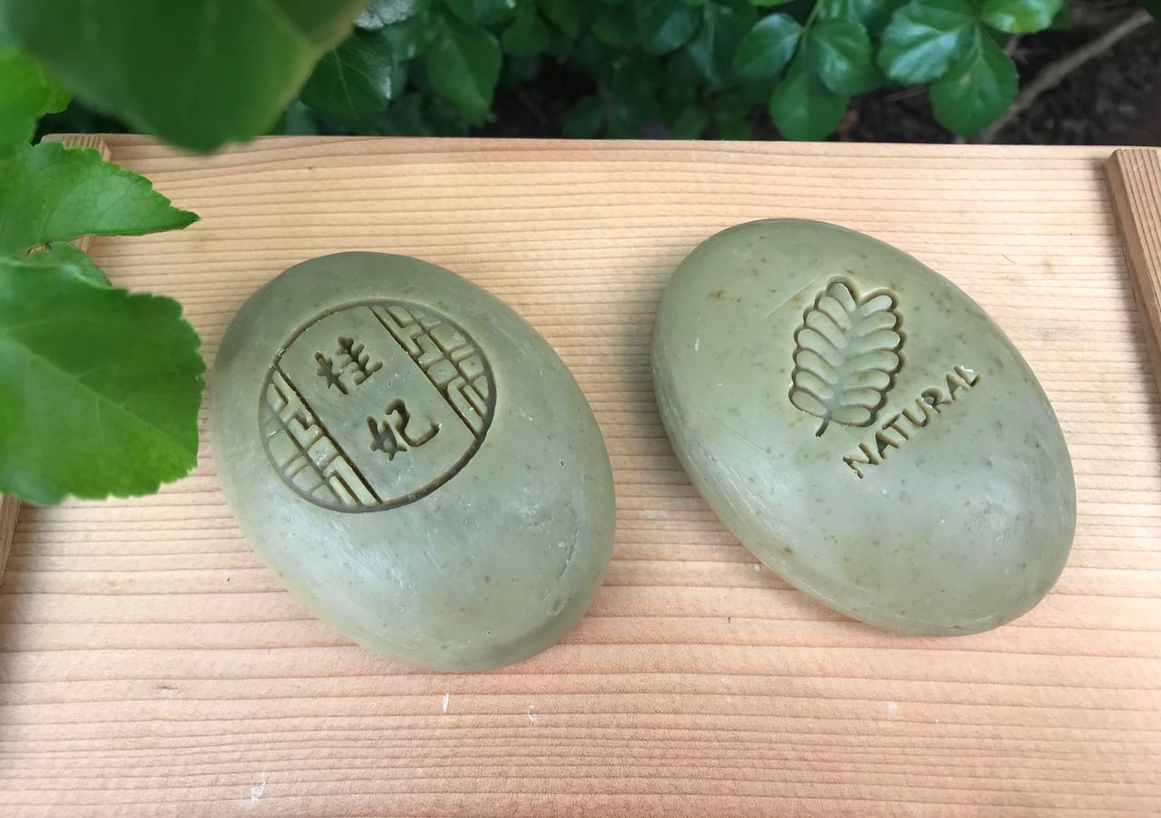 Two smooth stones with engraved designs: one with a leafy branch and the word "NATURAL," the other with Chinese characters and a decorative border, placed on a wooden surface with green foliage in the background.