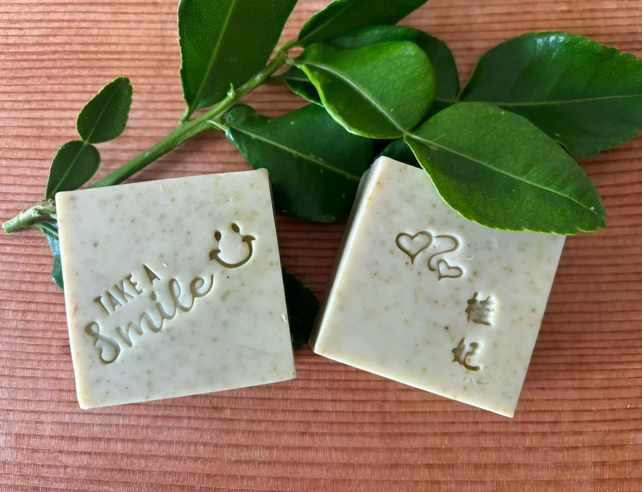 Two sheets of soap with engraved messages, one saying 'Take a smile' with a smiley face and the other with heart symbols and Korean characters, placed on a textured pink surface with green leaves.