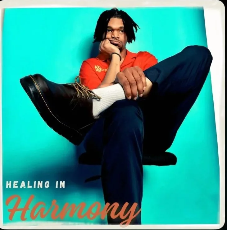 A young man with dreadlocks sitting in a relaxed pose, wearing a red shirt, navy pants, and black shoes, against a blue background. Text says "Healing in Harmony."