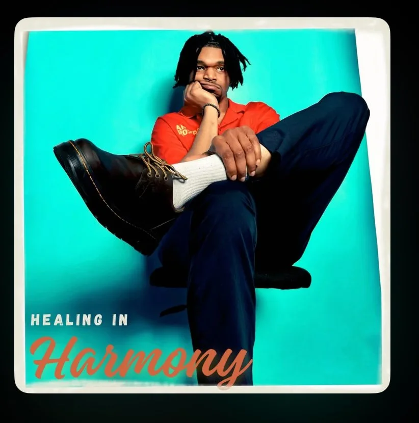 with dreadlocks wearing a red shirt, sitting on a chair with one leg crossed over the other, against a bright blue background. Their hand is resting on their chin, and they are wearing black boots, white socks, and dark pants. The text on the image says 'Healing in Harmony'.