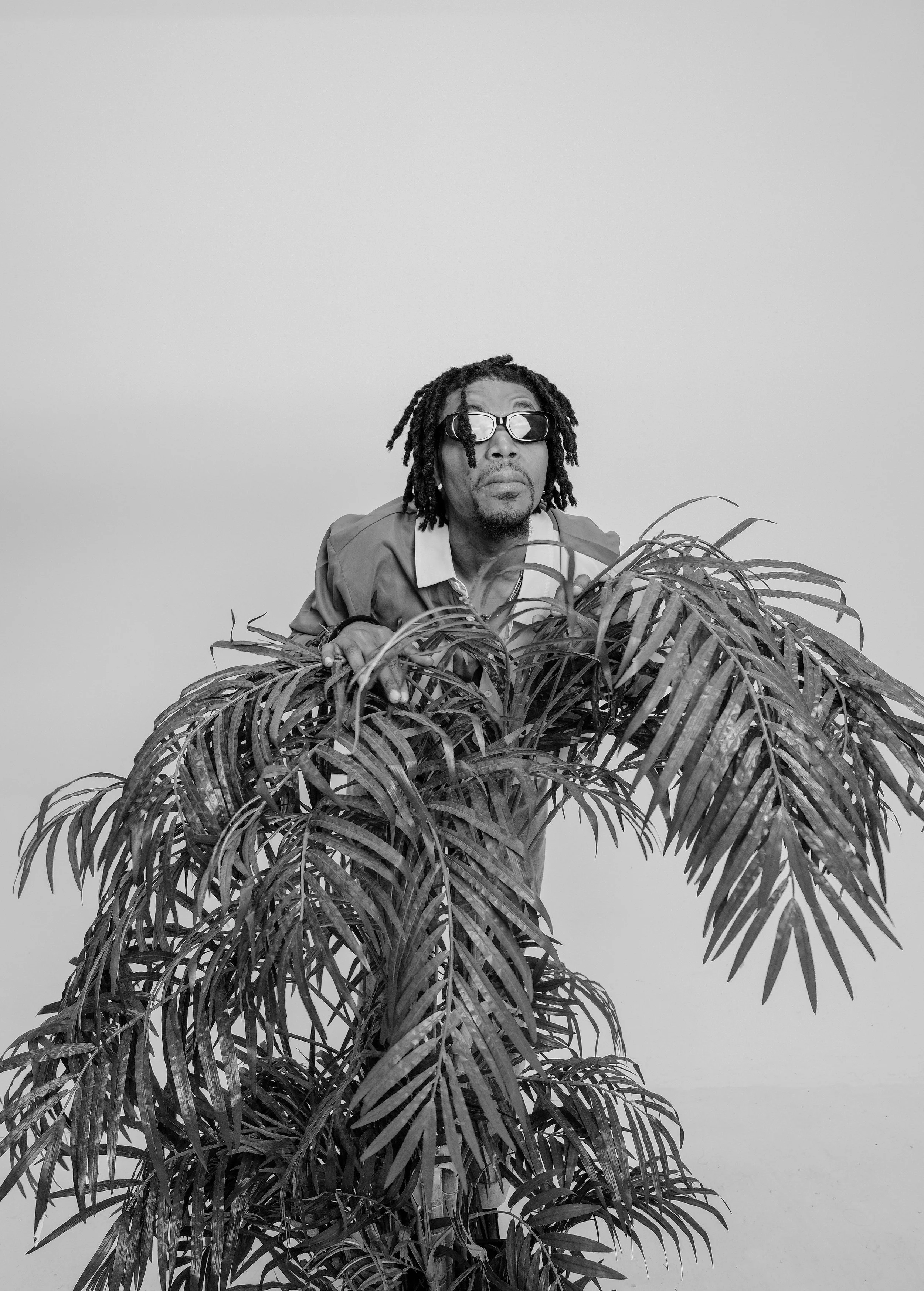 A man with Dreadlocks wearing glasses and a jacket, leaning over a large leafy plant against a plain background.