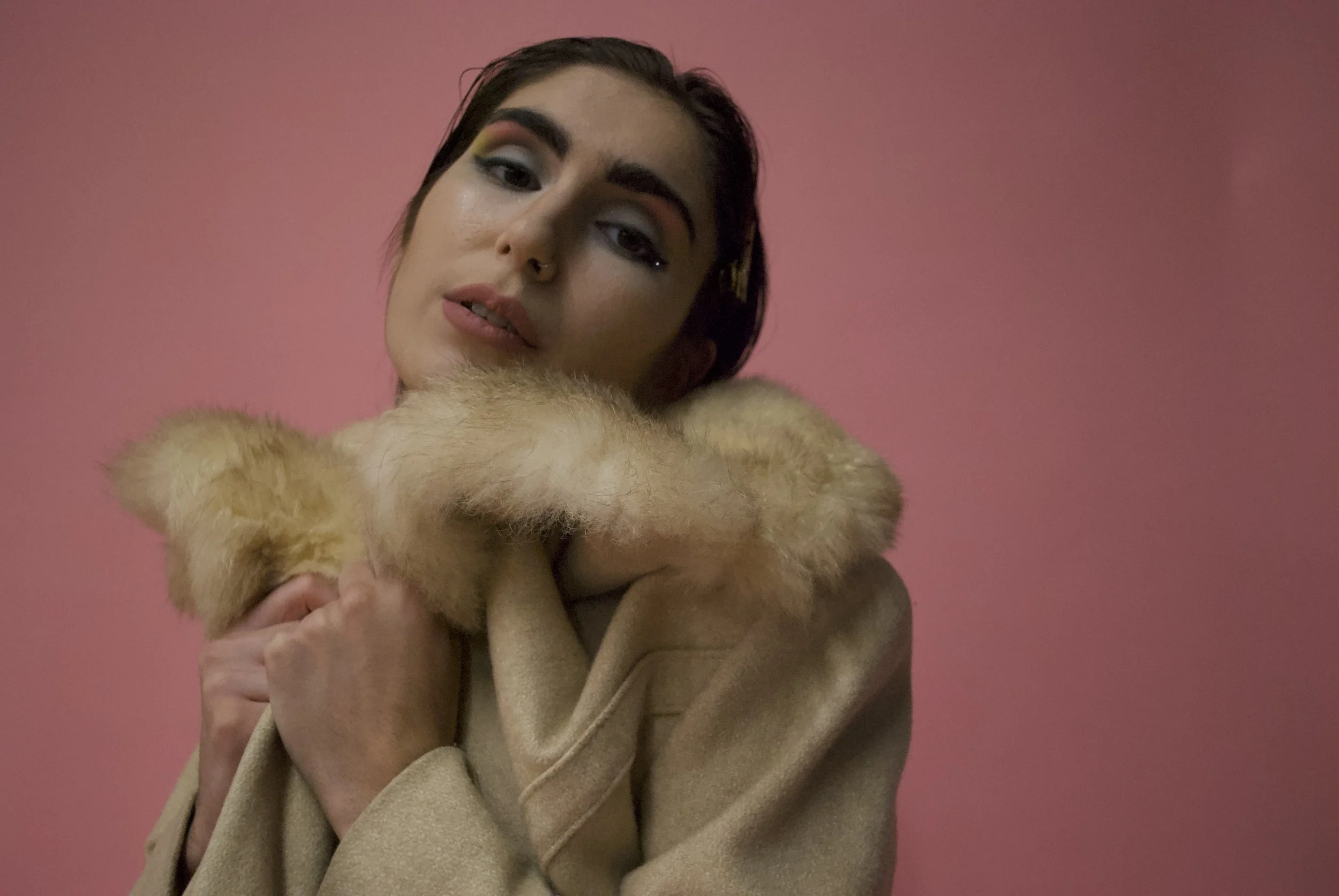 A woman with dark hair and colorful eye makeup holding a tan fur coat against a pink background.