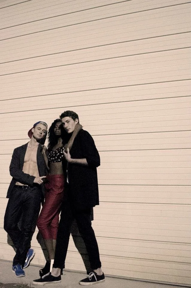 Three young people standing together in front of a large beige wall with horizontal lines. The person on the left has purple hair, a cap, an open blazer, and no shirt underneath. The person in the middle has curly black hair, a black polka dot crop top, and red pants. The person on the right has short hair, a black long shirt or jacket, and black pants, with their arm around the person in the middle.