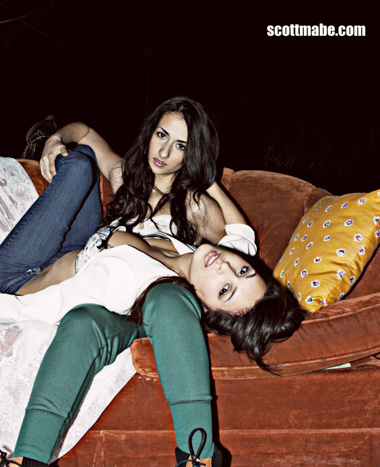 Two young women lying on a brown couch, with one resting her head on a pillow and the other sitting upright, both looking at the camera.