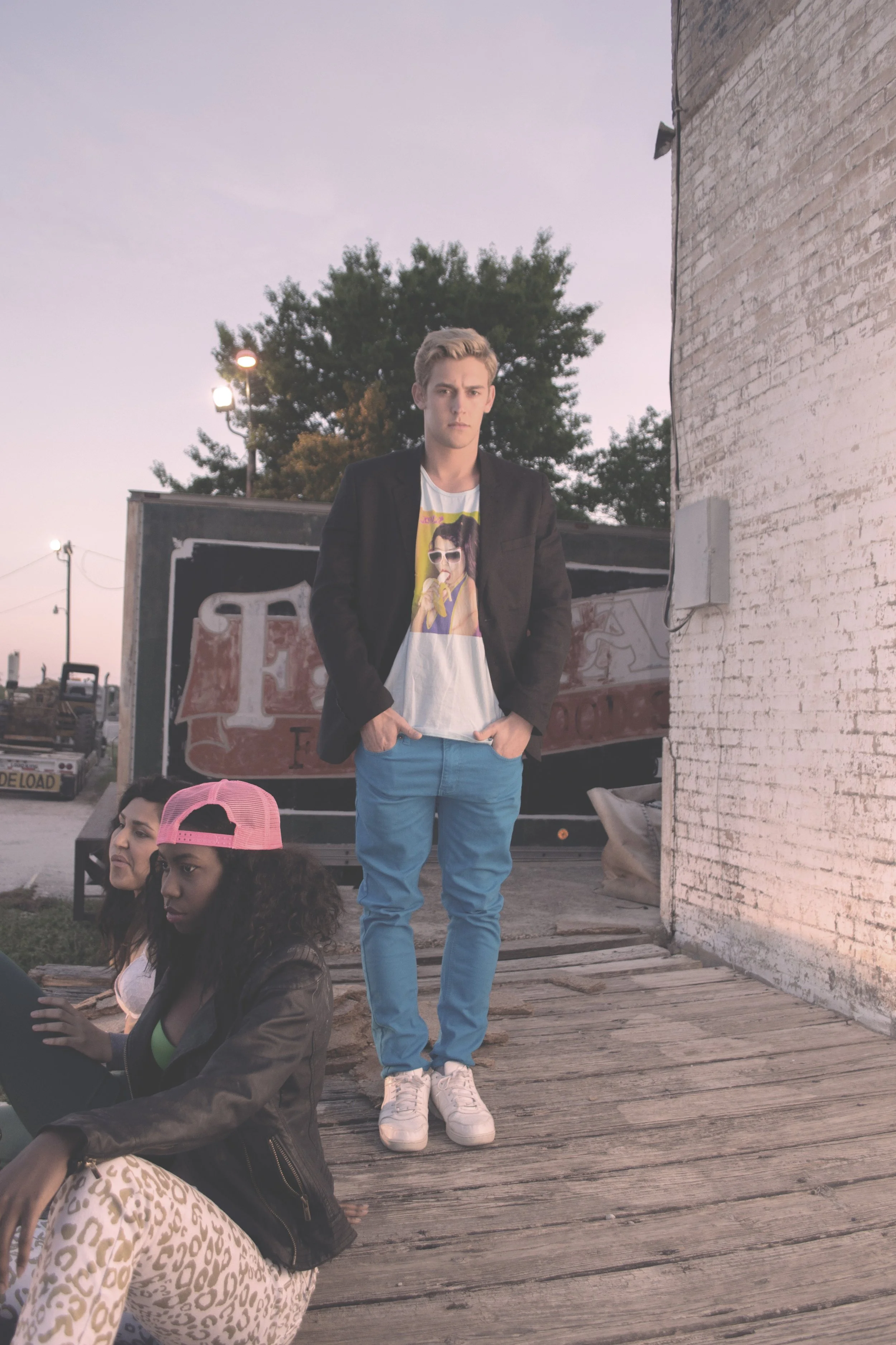 Three young people outdoors during sunset; one standing man wearing a black jacket, graphic t-shirt, bright blue pants, and white sneakers, with hands in pockets; two women sitting on wooden deck, one in leopard print pants and black leather jacket, and the other with a pink cap and dark jacket, all near an old building and trailer.