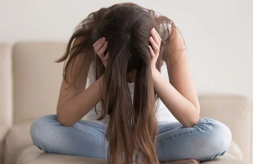 A teen girl sitting on a sofa with her arms covering her face and her long hair hanging down, holding her head in her hands.