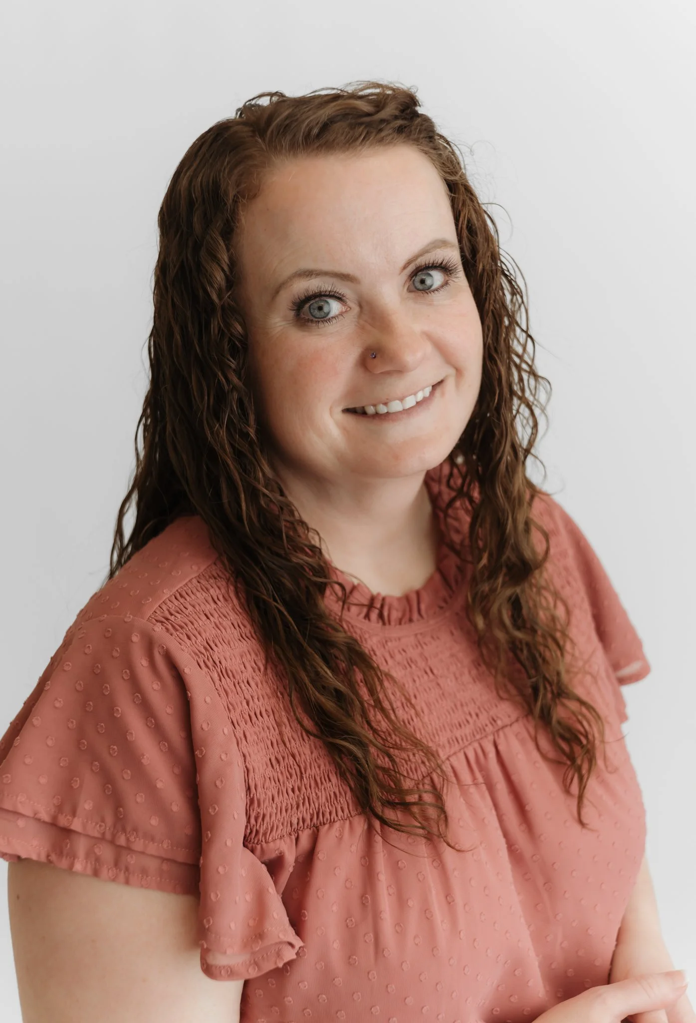 Close-up of a woman with long, curly red hair, blue eyes, and a nose piercing, wearing a pink textured blouse with puffed sleeves, smiling softly against a plain white background.