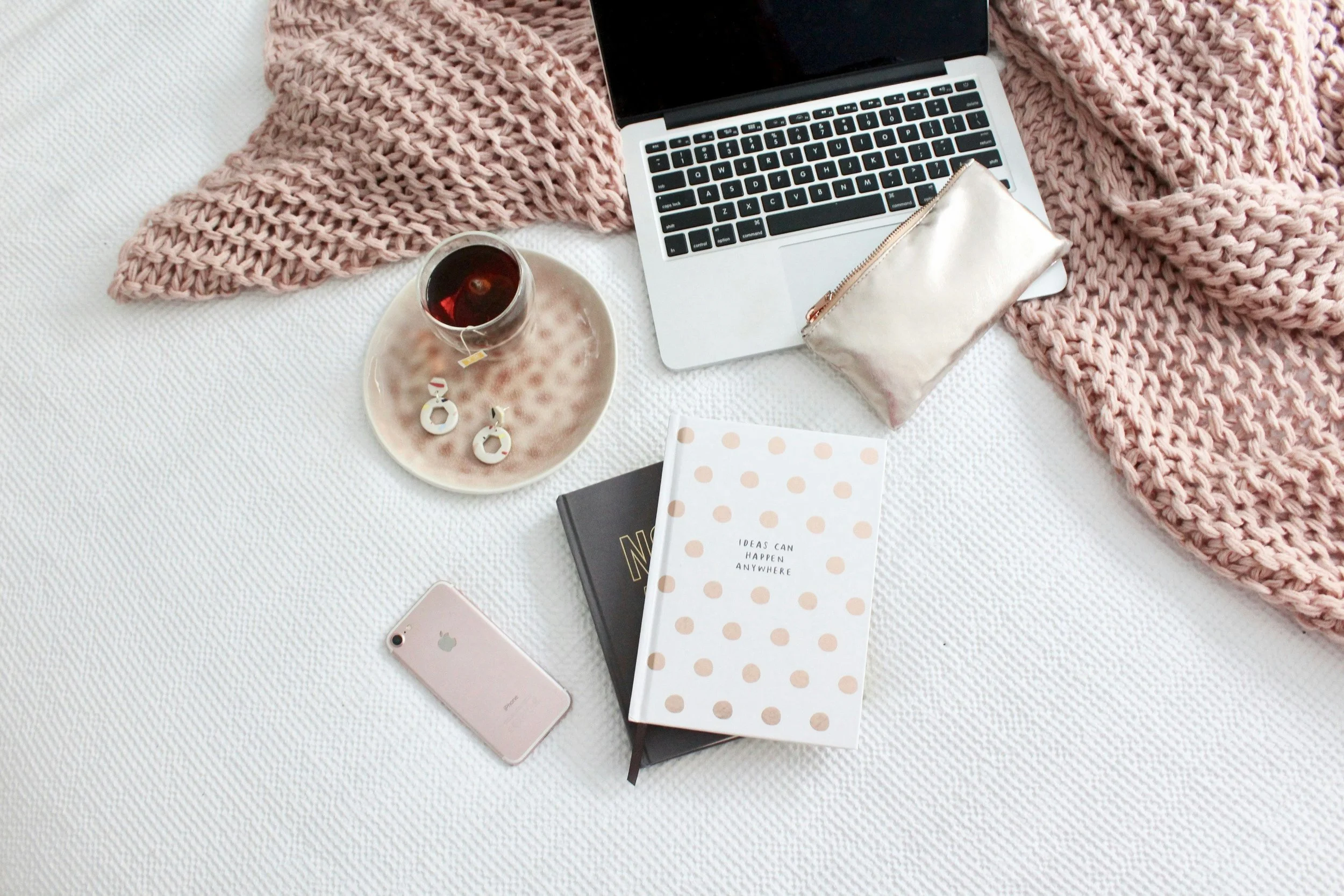 A flat lay of a cozy workspace with a laptop, pink chunky knit blanket, a cup of tea with a tea bag, a pair of white earrings, a rose gold pouch, a pink iPhone, a black notebook, and a polka dot planner with the text "Ideas can happen anywhere" on a white textured surface.