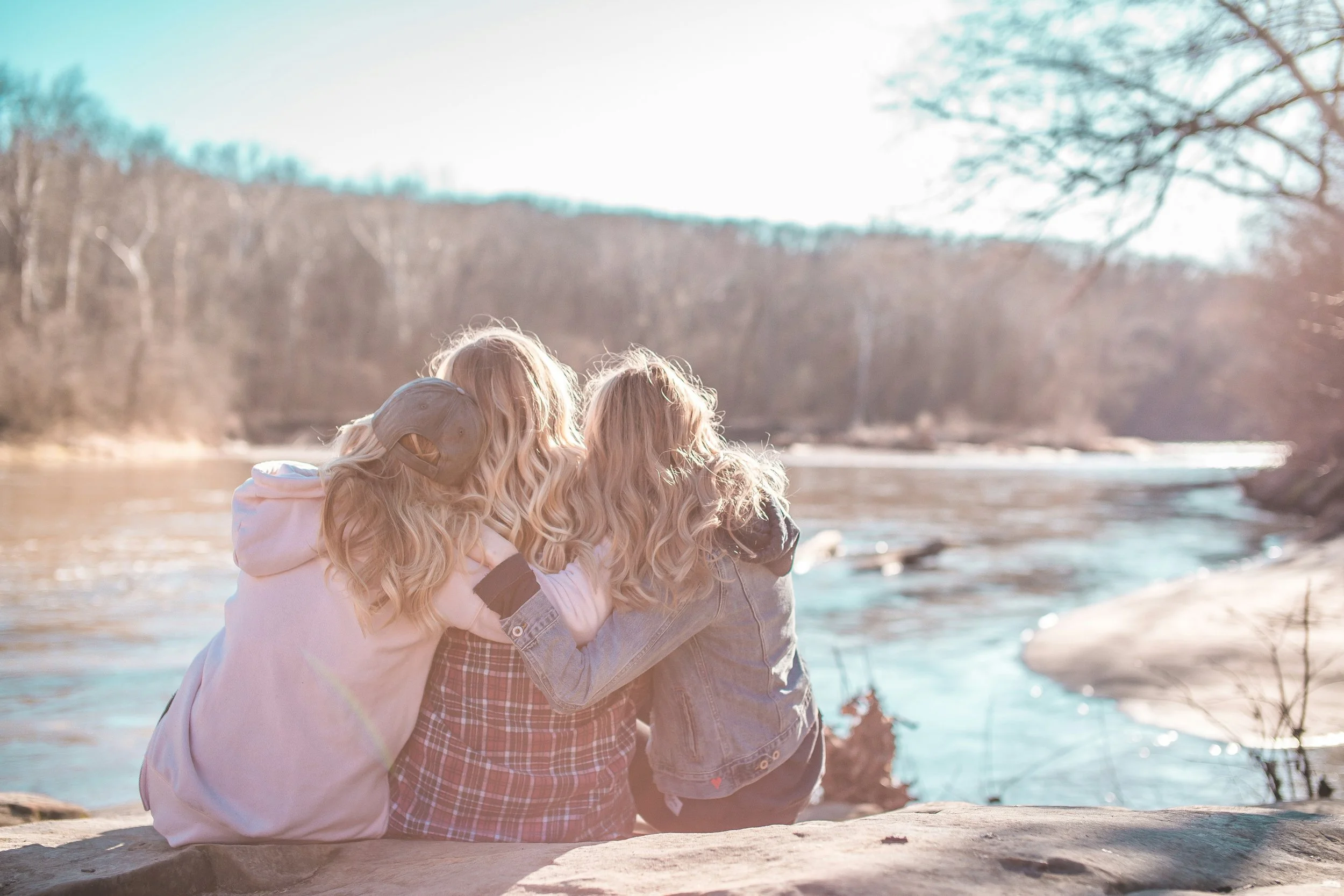 Three teen girls embracing and sitting on a rock by a river, enjoying the view of trees and hills in the background.