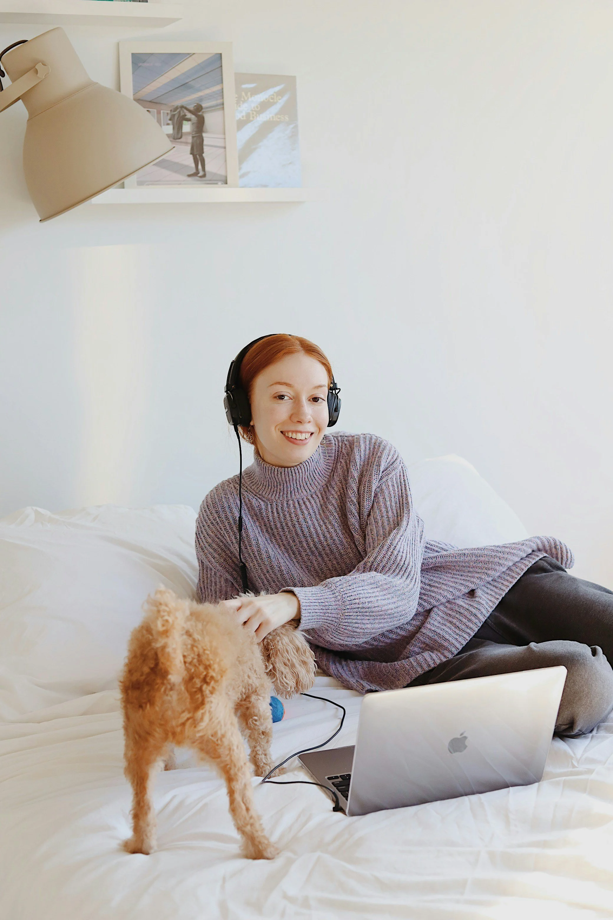 A teen girl lying on a bed with a small fluffy dog, wearing headphones, using a laptop, in a bright room with pictures on the wall.