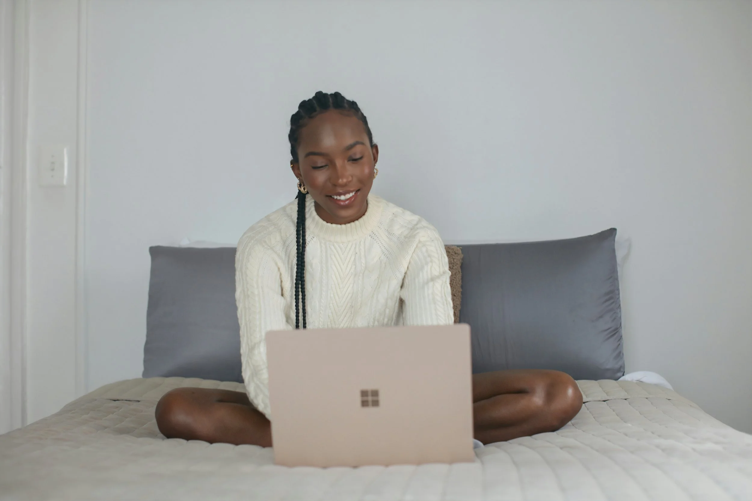 Teen girl booking a 1:1 confidence coaching session sitting cross-legged on a bed with grey pillows, using a laptop, smiling, in a minimalistic bedroom with a plain white wall.