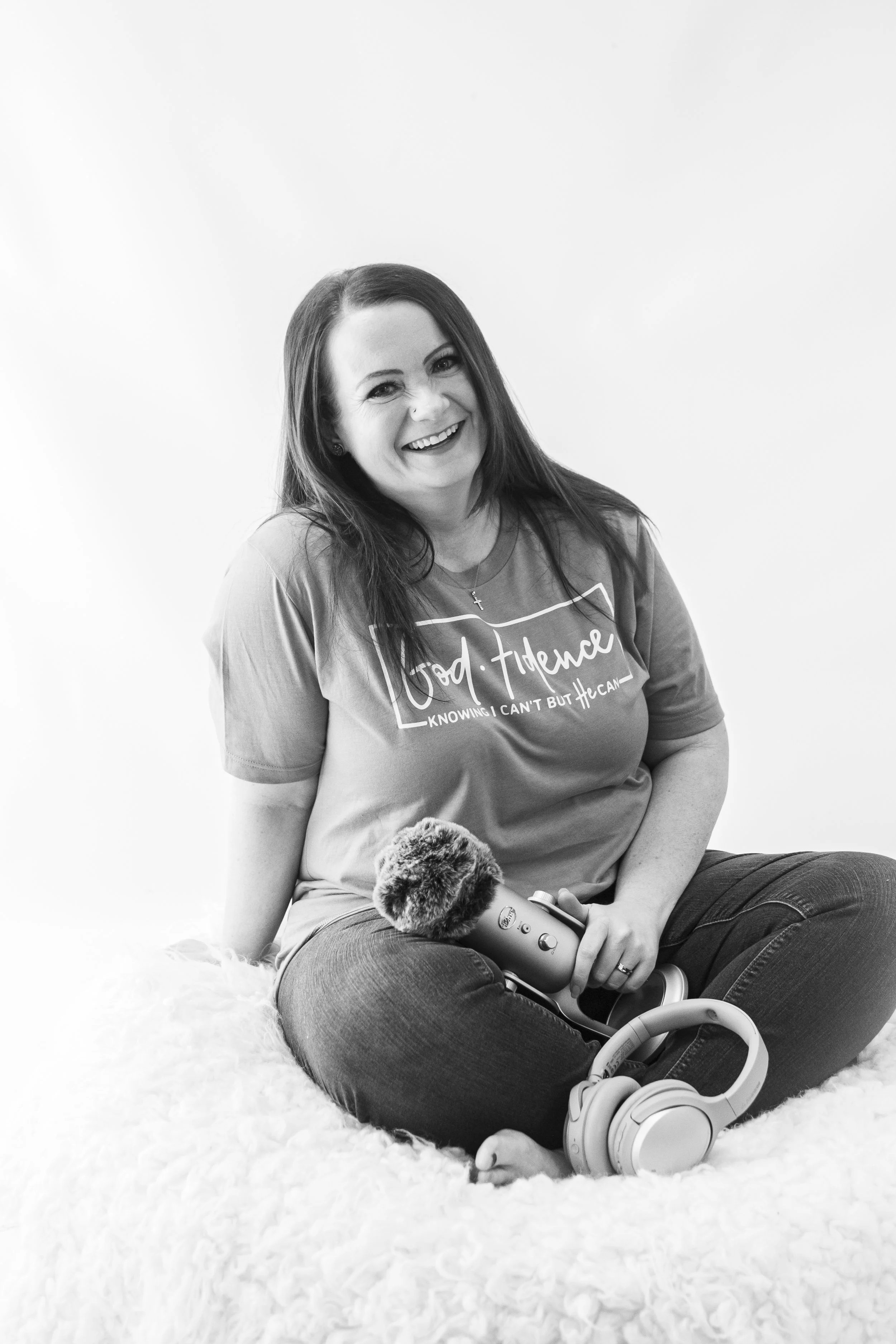 A woman sits on a fluffy surface, smiling and holding a microphone, with a pair of headphones resting on her knee.