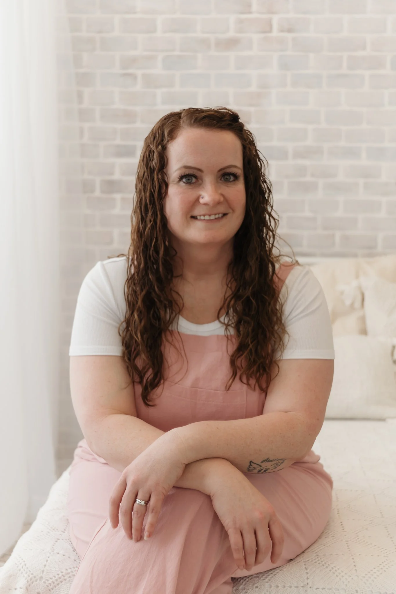 A woman with long, curly brown hair, blue eyes, and a nose piercing sitting on a bed against a white brick wall. She is smiling, wearing a white top and pink overalls, with crossed arms and a tattoo on her forearm.