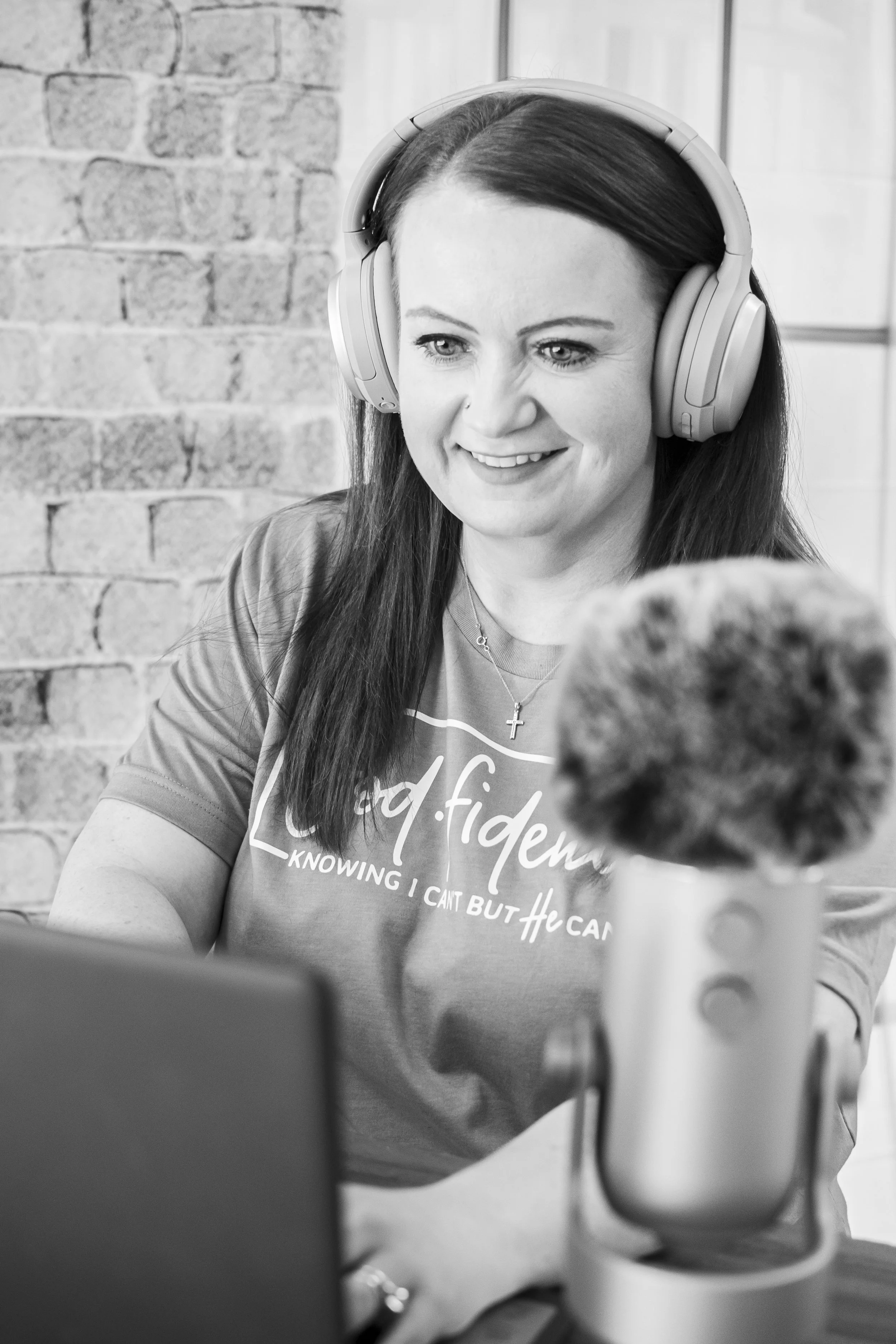 A woman wearing headphones smiling at a microphone in a recording or podcasting studio with a brick wall in the background.